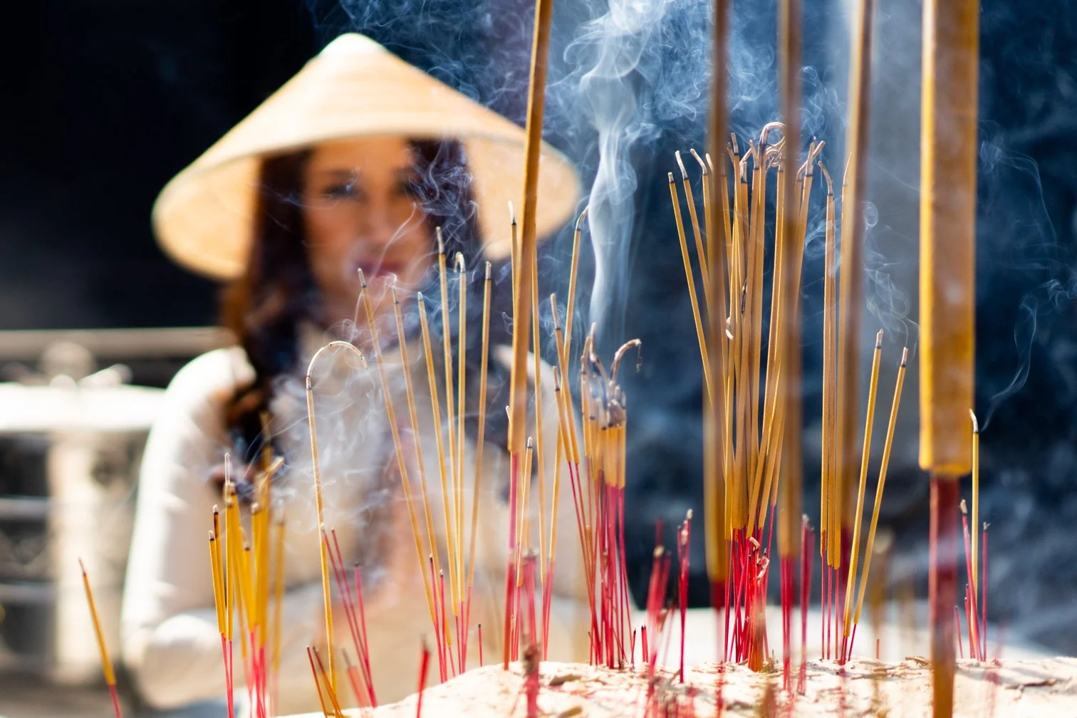A woman wearing a traditional Asian straw hat is in the background, slightly out of focus, with a large plate of incense sticks in the foreground burning and emitting smoke.
