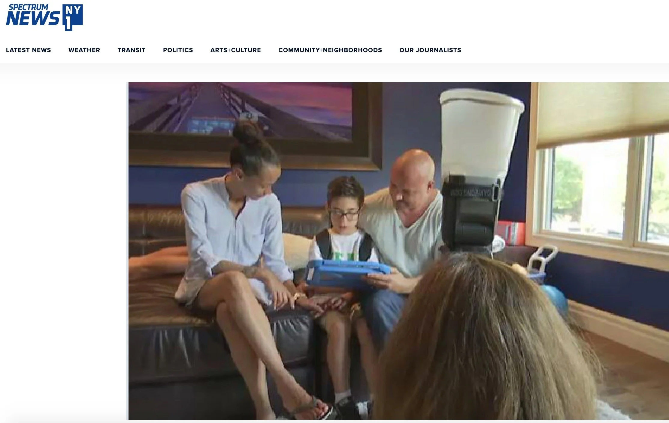 A family sitting on a leather couch in a room, looking at a device together. Two adults and one child with glasses, with a window and a large lamp in the background.