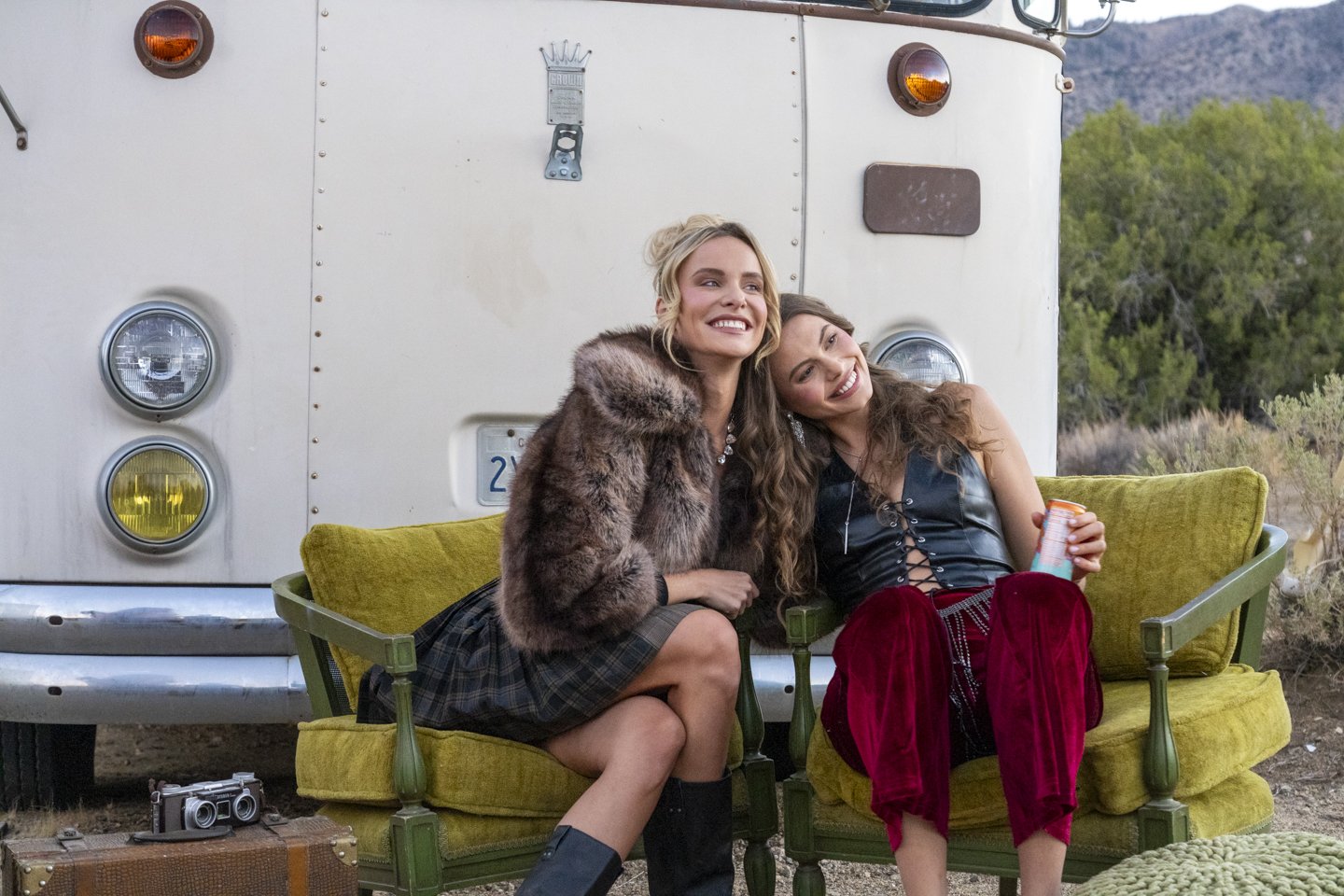 Two women sitting on a vintage green couch in front of a white van, smiling and enjoying each other's company, with a camera on a suitcase nearby and a scenic outdoor background.