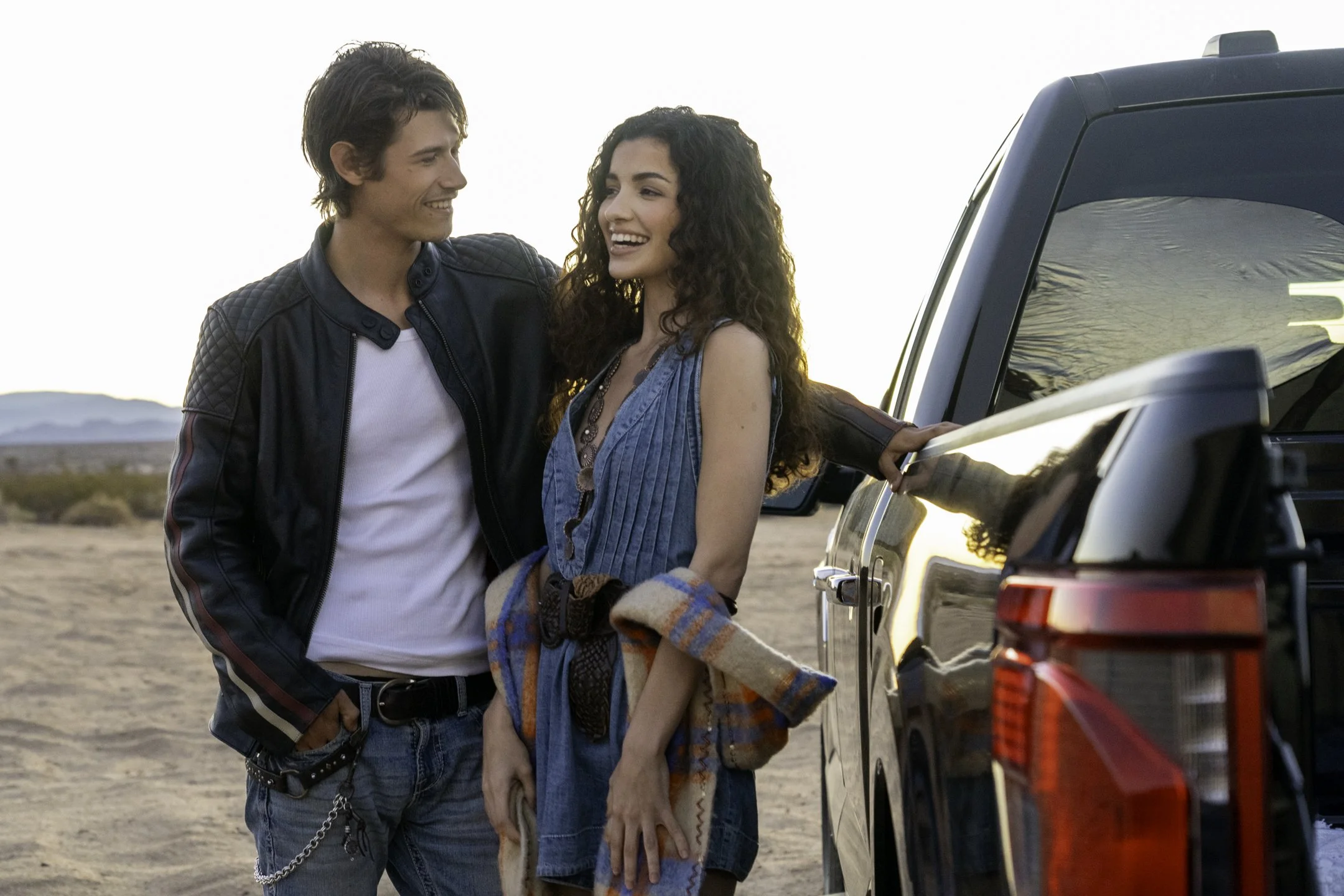A young man and woman stand next to a black pickup truck in a desert landscape during sunset, smiling and looking at each other.