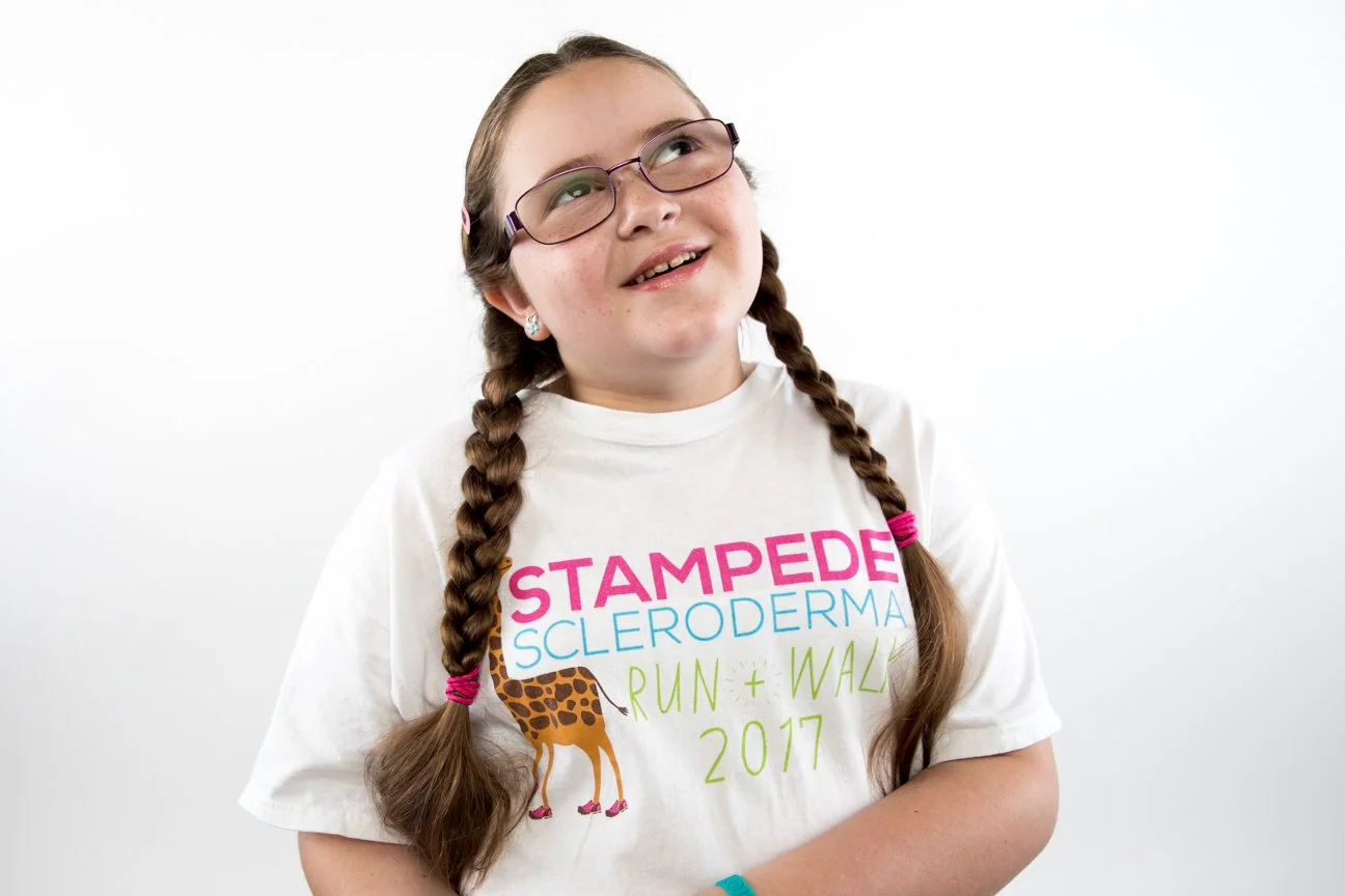 A young girl with glasses and long braided hair wearing a white T-shirt with the words 'StampeDe Scleroderma Run + Walk 2017' and a giraffe graphic, smiling and looking upward.
