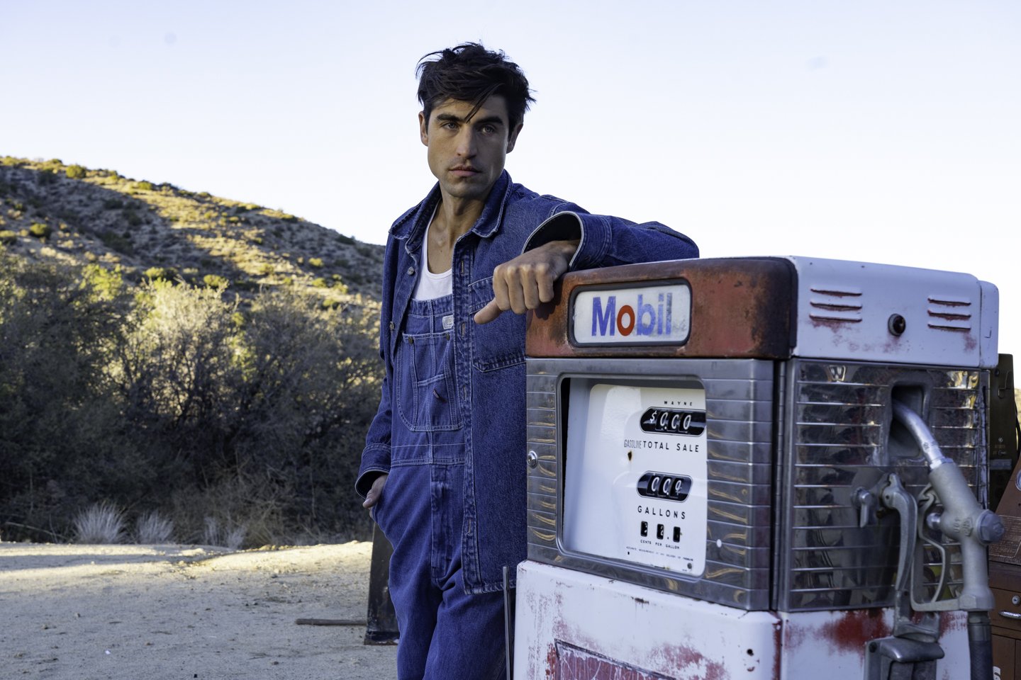 A young man in a denim jacket standing next to an old Mobil gas pump in a desert landscape.