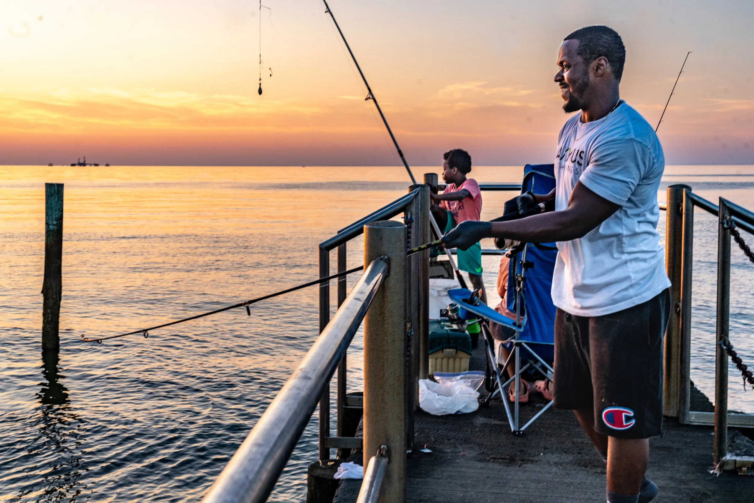 People fishing on a pier during sunset, with the ocean and a ship in the distant background.