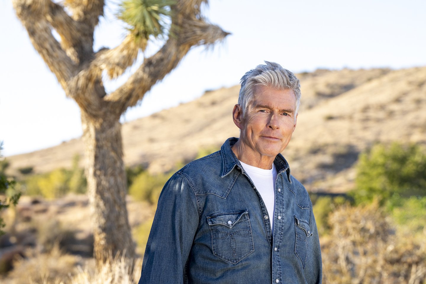 A mature man with gray hair wearing a denim jacket outdoors in a desert landscape with a Joshua tree in the background.