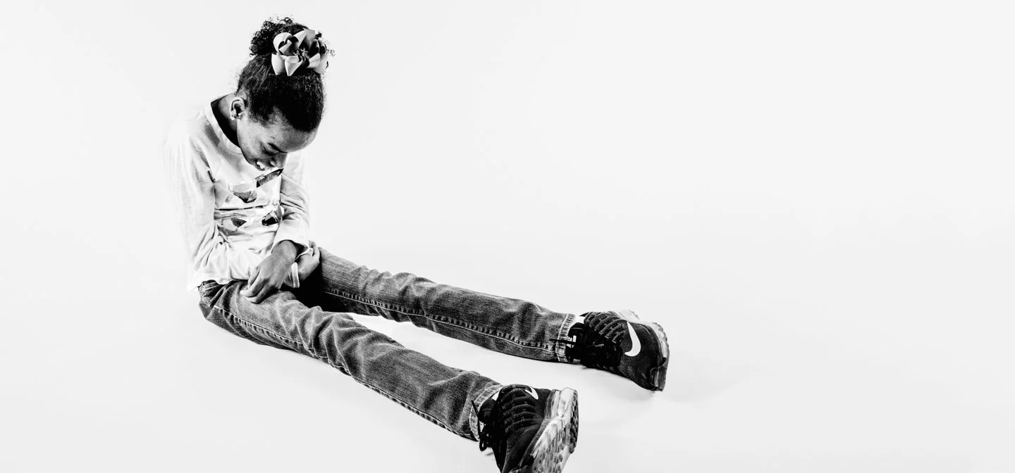 A young girl sitting on the floor, laughing, wearing a long-sleeved shirt, jeans, sneakers, and a bow in her hair, against a plain white background.