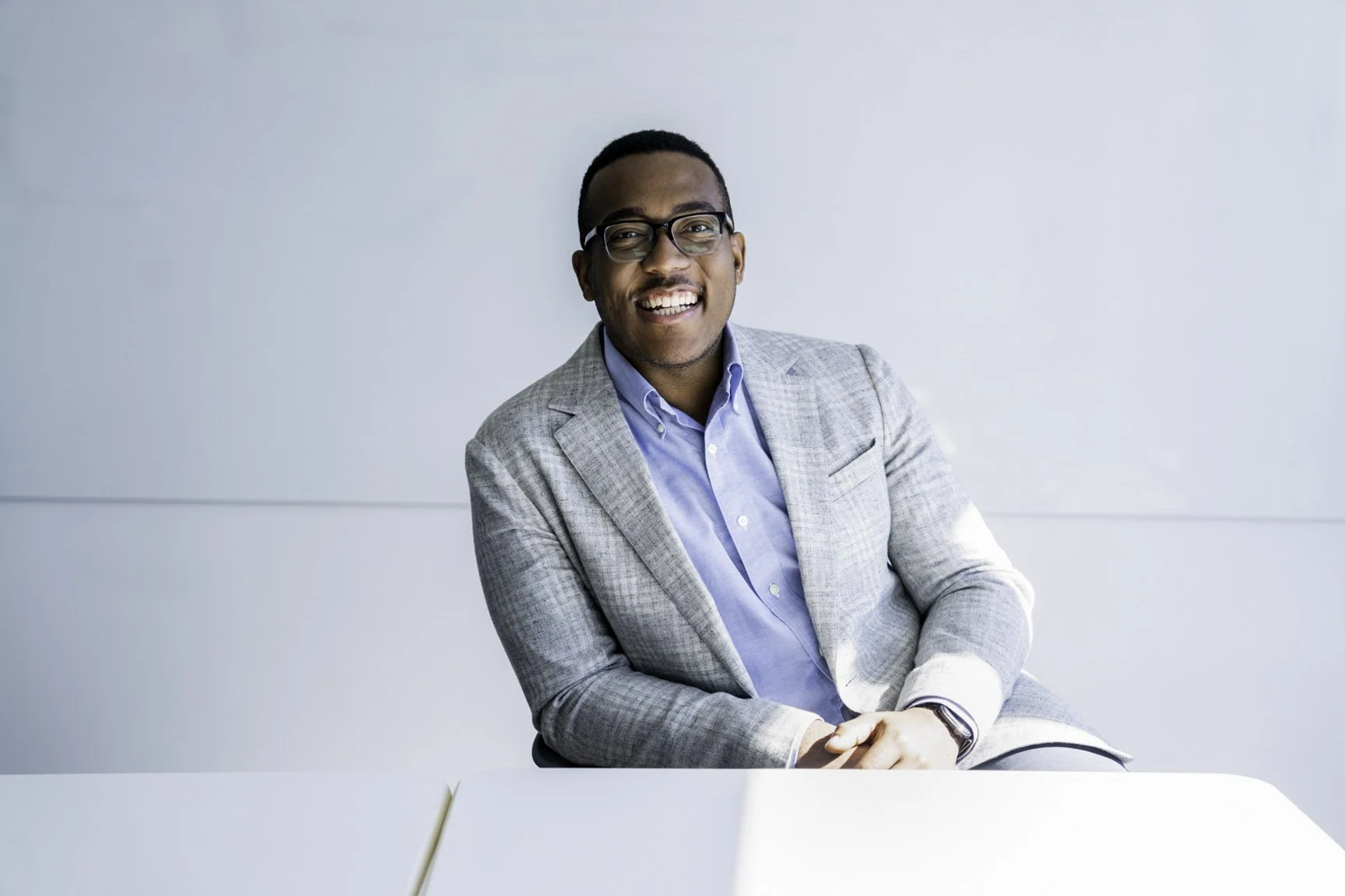 A man wearing glasses, a light gray blazer, and a blue shirt sitting at a white table, smiling in front of a plain light-colored wall.