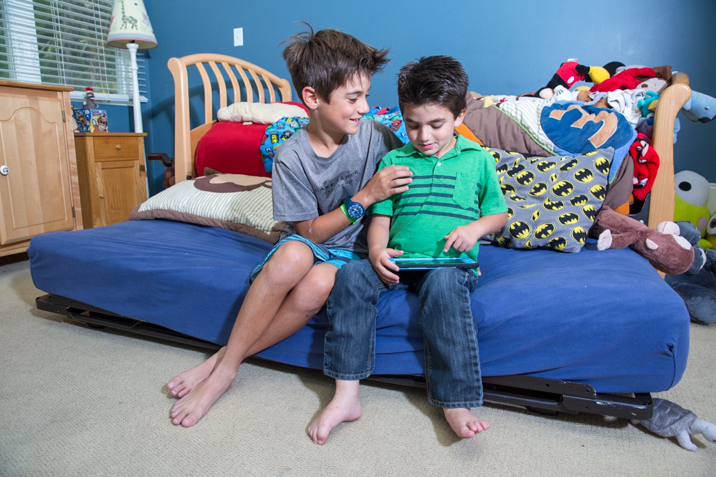 Two boys sitting on a bed, playing on a tablet, surrounded by pillows and stuffed toys, in a child's bedroom with blue walls.