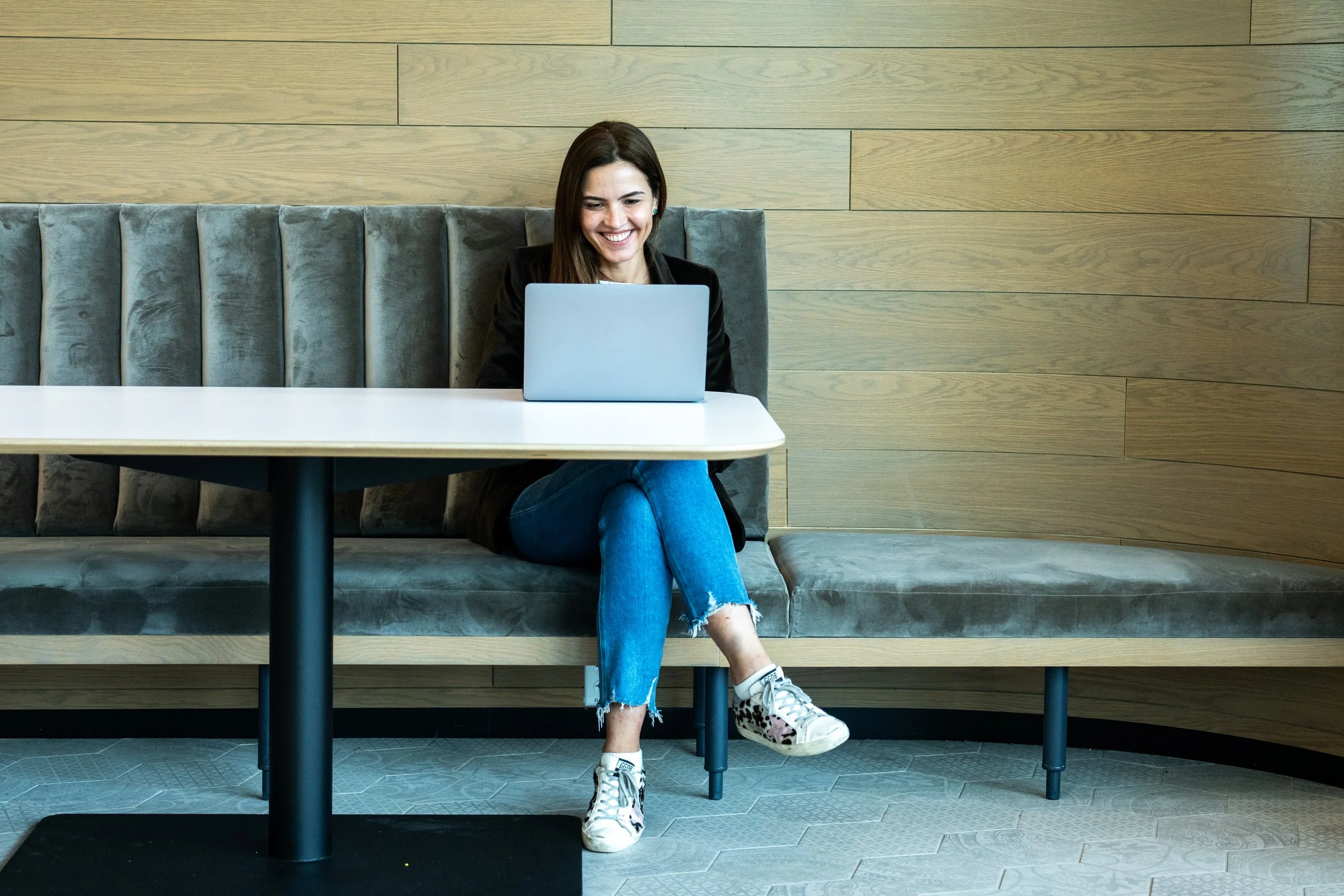 Young woman sitting on a gray upholstered bench, smiling at her laptop in a modern wooden interior.