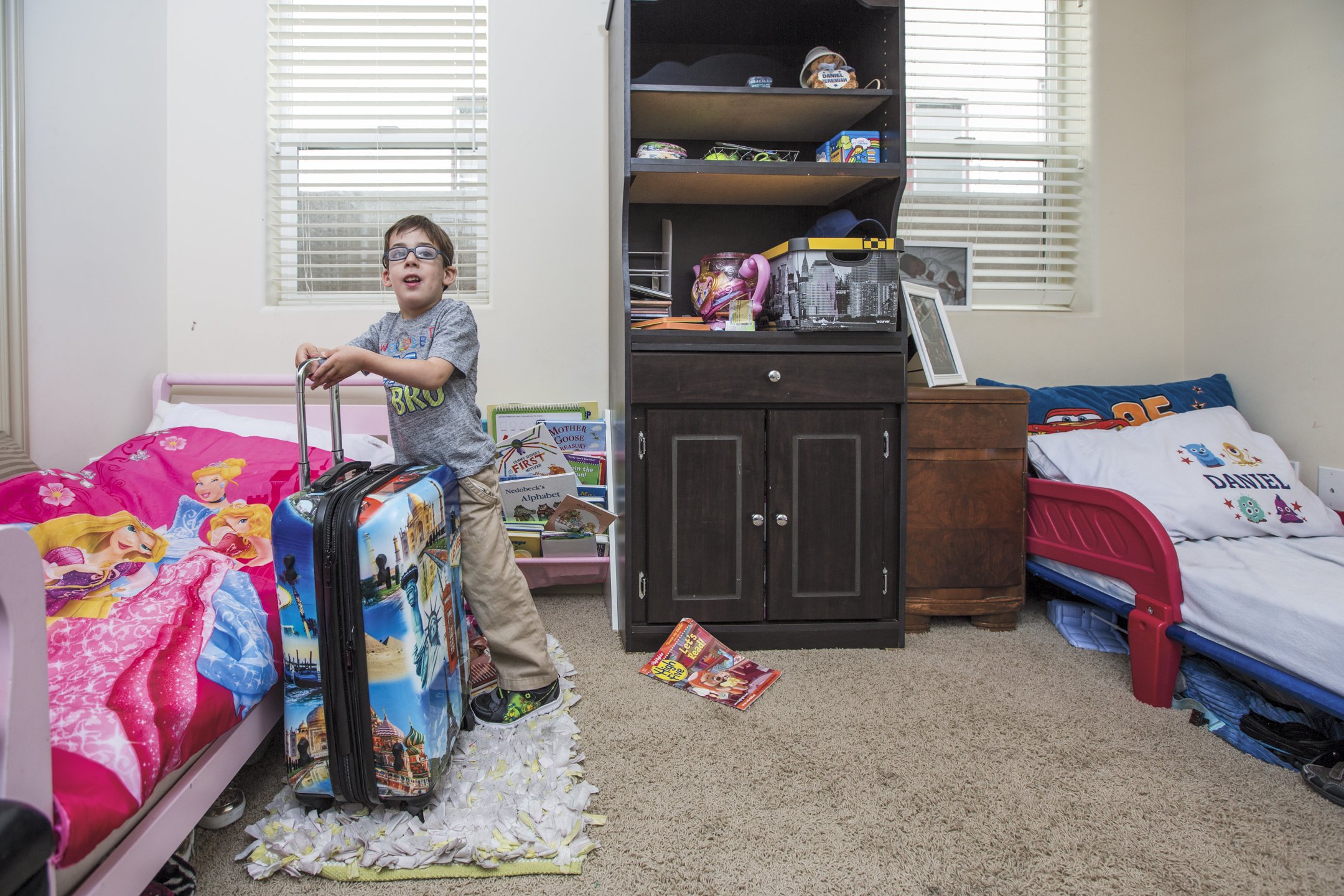 A young boy in glasses standing next to a pink bed with Disney princess sheets, holding a suitcase in a child's bedroom with stuffed animals, books, and a bookshelf.