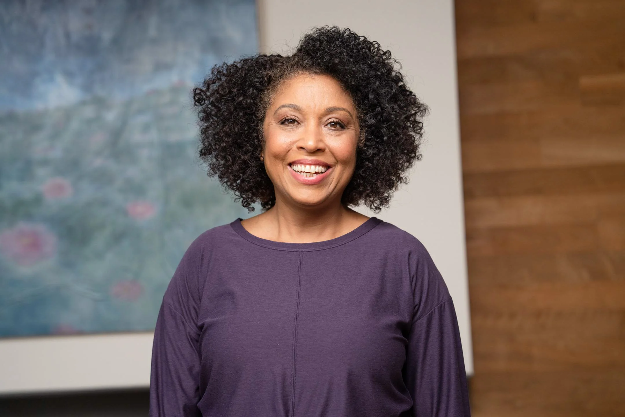 Smiling woman with curly black hair wearing a purple shirt standing indoors with abstract art and wooden wall in background.