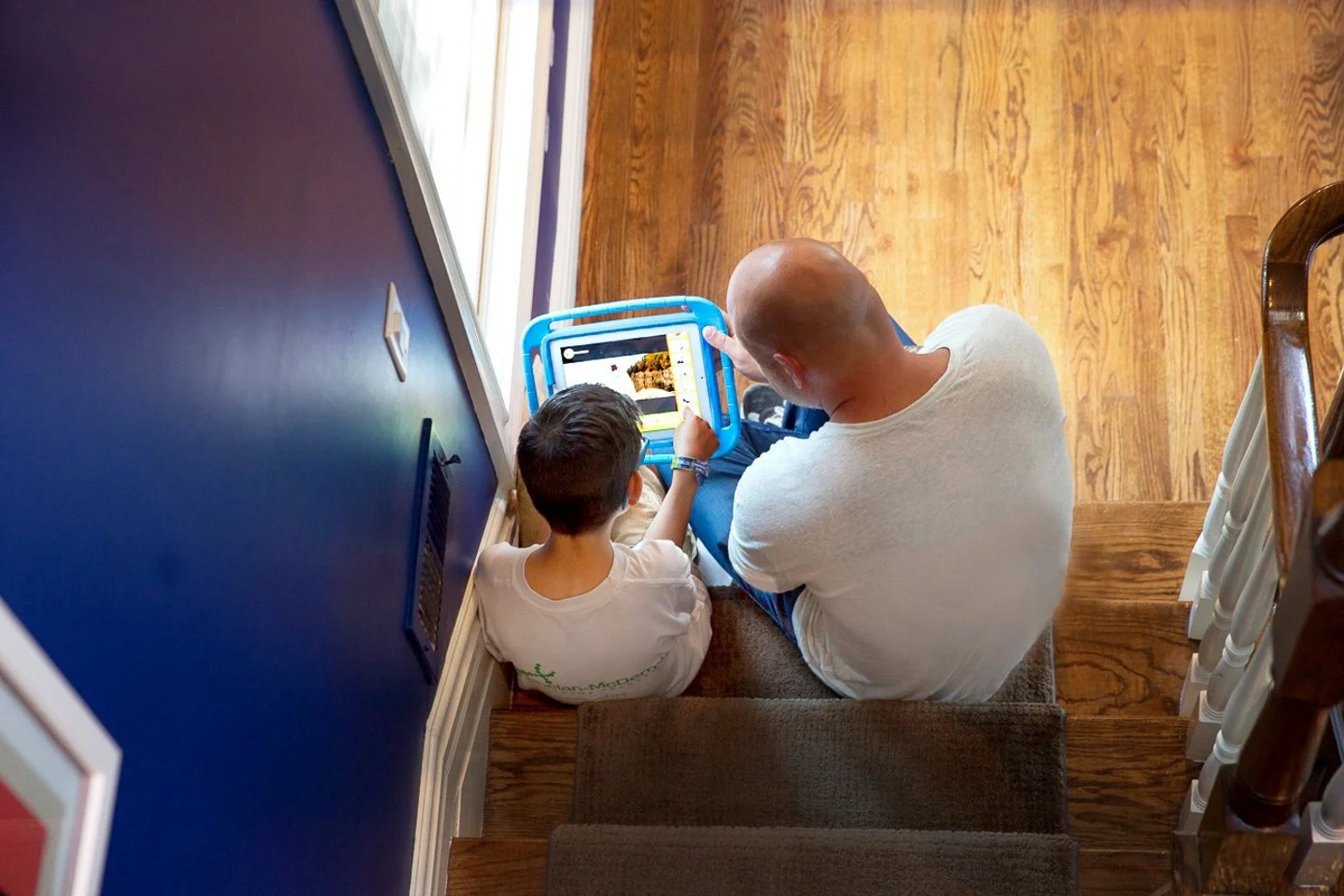 An adult and a child sitting on the stairs, watching a tablet together, with a blue railing nearby and hardwood floors.