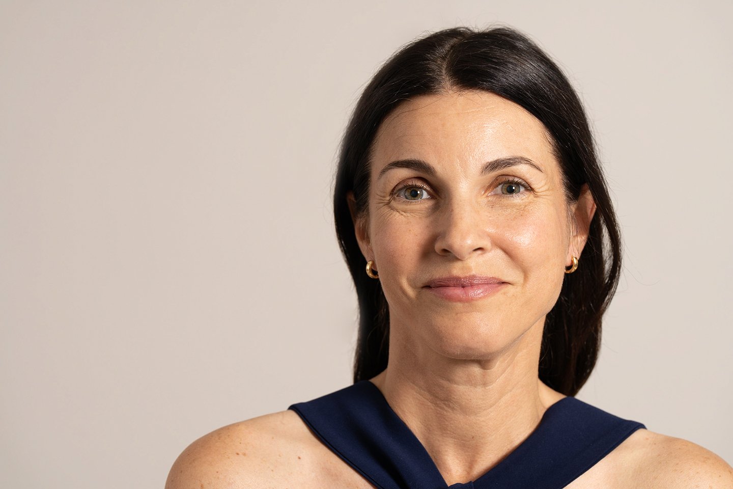 A woman with dark hair and light skin, wearing a dark blue sleeveless top and small gold hoop earrings, smiling subtly against a plain light background.
