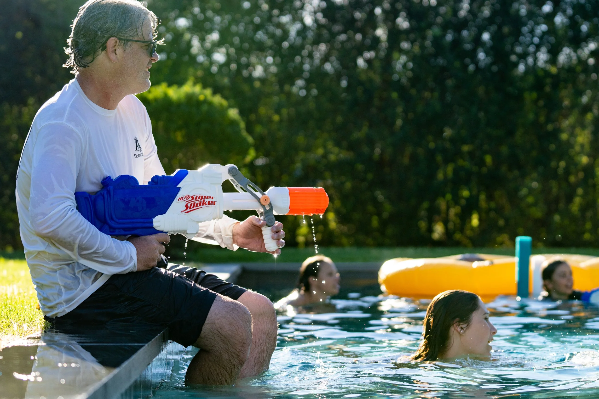 A man sitting on the edge of a pool, wearing sunglasses and a white shirt, holding a Nerf Super Soaker water gun, while several people swim in the pool nearby on a sunny day with green trees in the background.