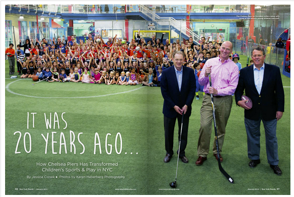 Large group of children and adults gathered in an indoor children's play area, with three men in the foreground holding hockey sticks and a football, celebrating an anniversary.