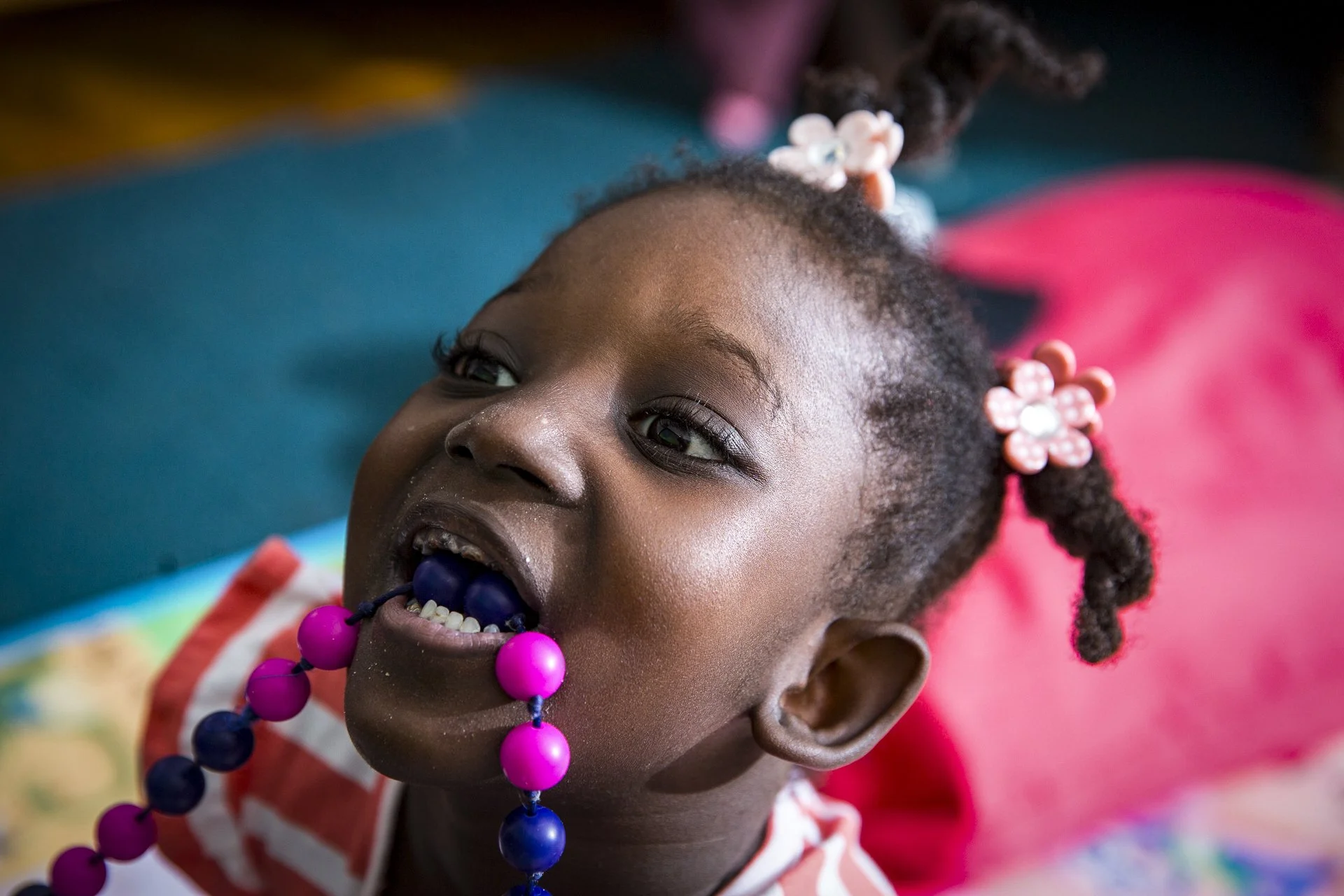 A young African girl with braided hair decorated with pink and white flower hair clips, lying on her back, smiling, with a colorful beaded necklace in her mouth.