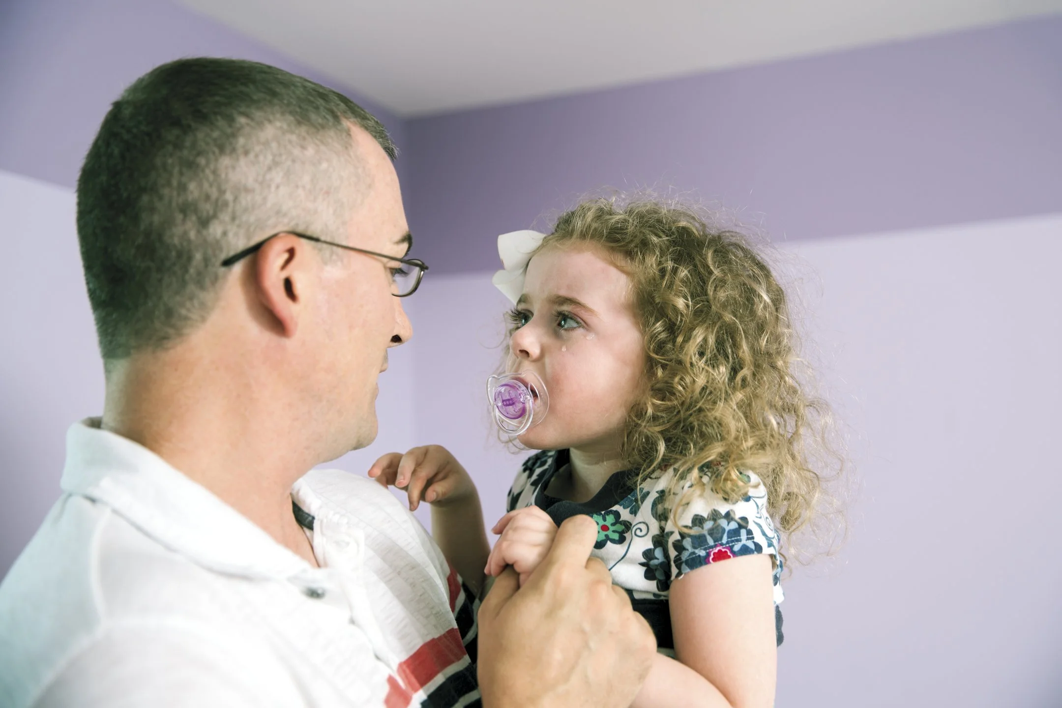 A man holding a young girl with curly hair and a pacifier in her mouth, looking at each other in a room with lavender walls.
