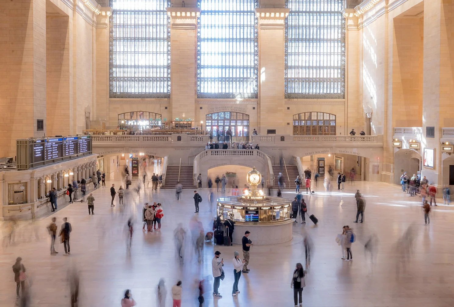 Grand interior of a large, historic train station with high glass windows, a central clock, and many people walking or waiting.