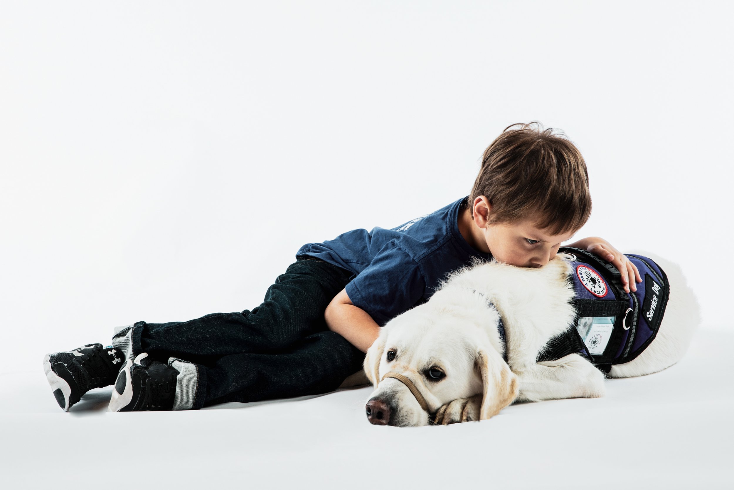 Boy lying on the floor next to a white Labrador puppy with a backpack, against a white background.