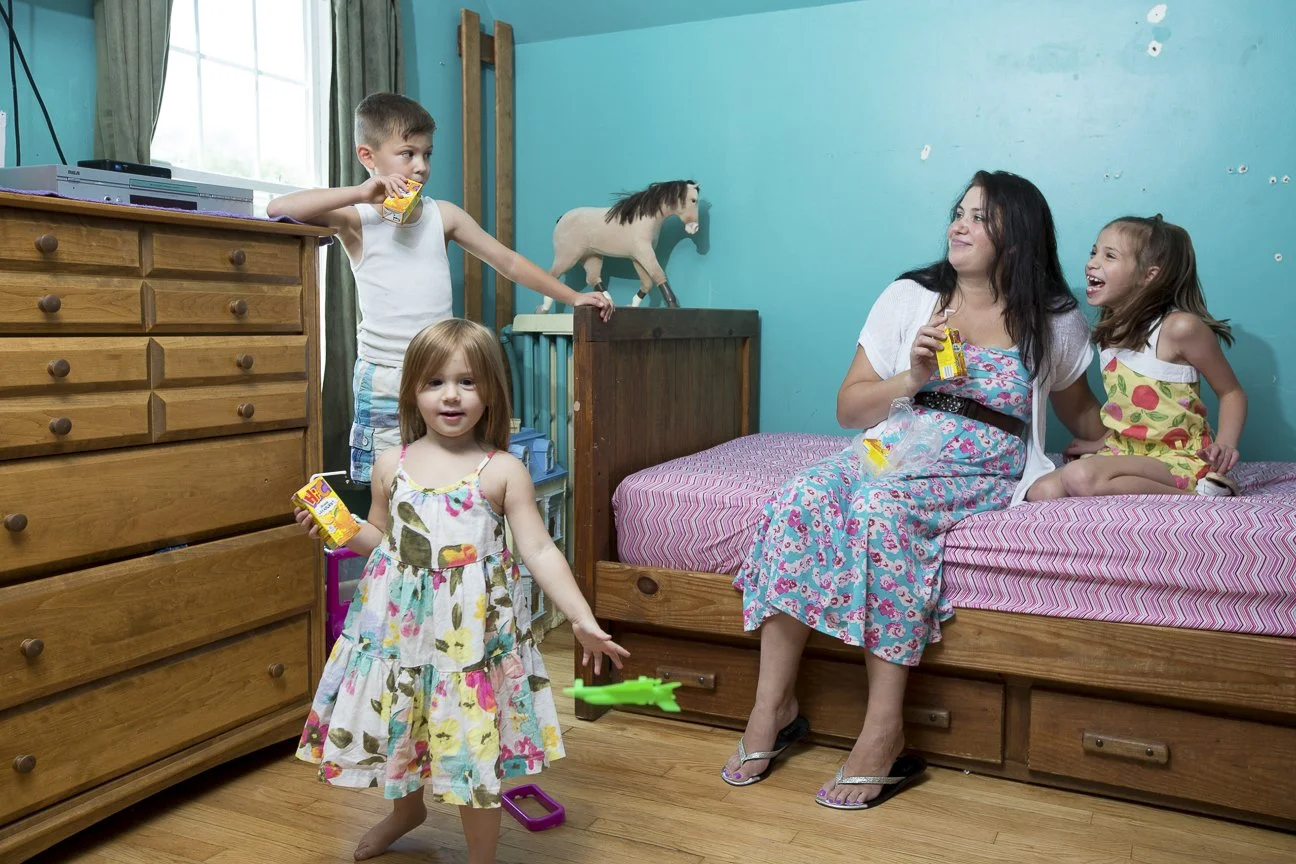 Four children and a woman sitting on a bed in a bedroom with teal walls and wooden furniture. One girl is standing with a juice box and Green toy, another girl and a boy are holding juice boxes, and the woman is also holding a juice box and smiling a