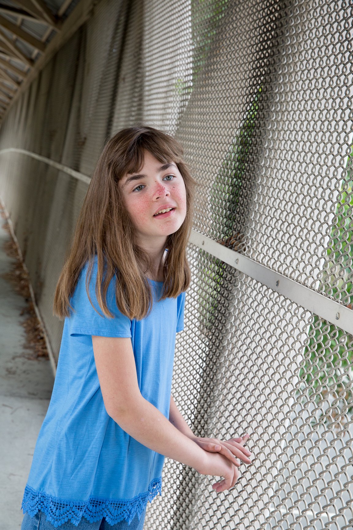 Young girl with long brown hair wearing a blue dress standing by a chain-link fence.