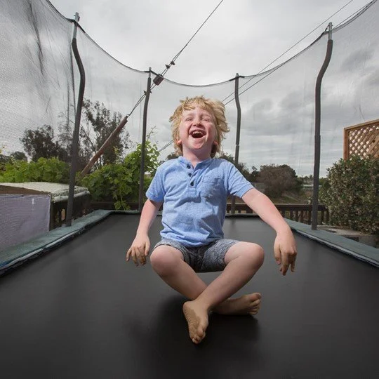 Child with curly blonde hair and a blue shirt sitting on a trampoline, laughing.