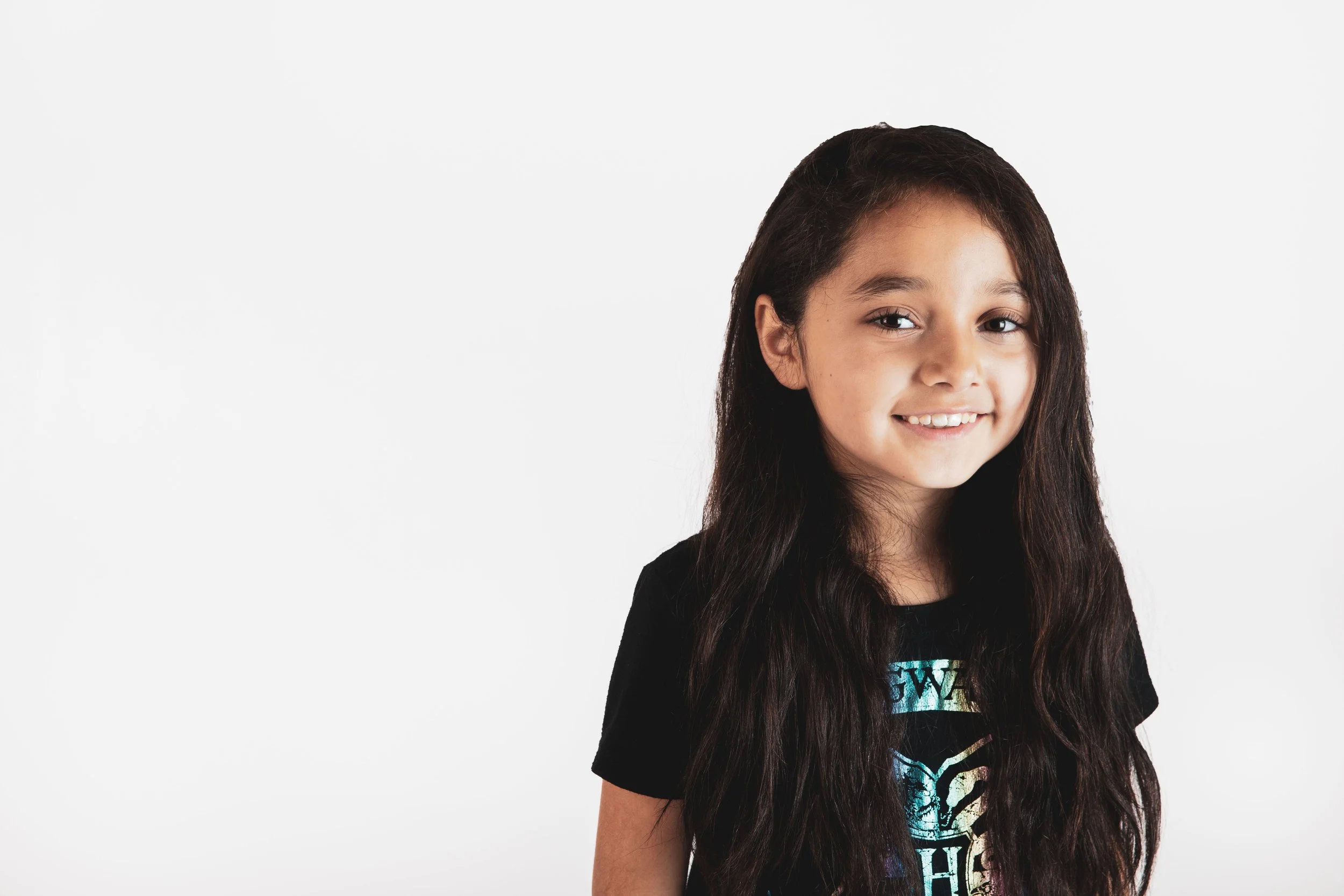 A young girl with long dark hair smiling, wearing a black t-shirt with a colorful graphic design, against a plain white background.