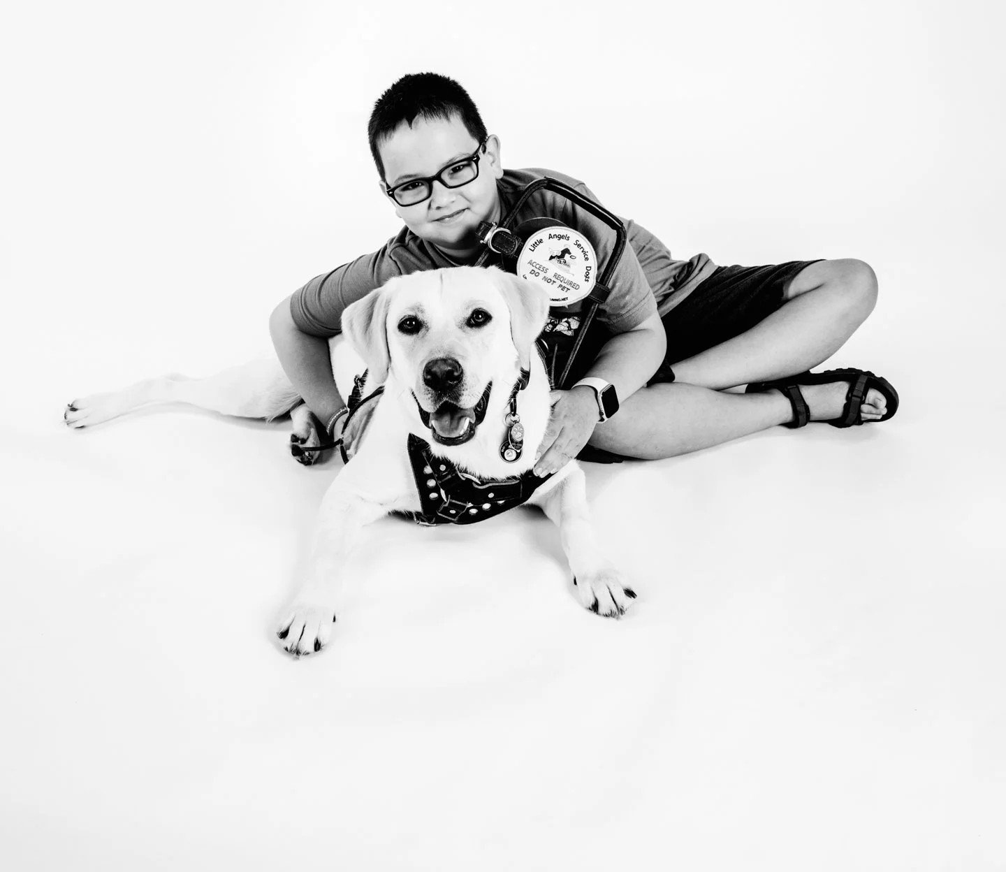 A boy with glasses and a smartwatch lying on the floor with a service dog, both smiling at the camera.