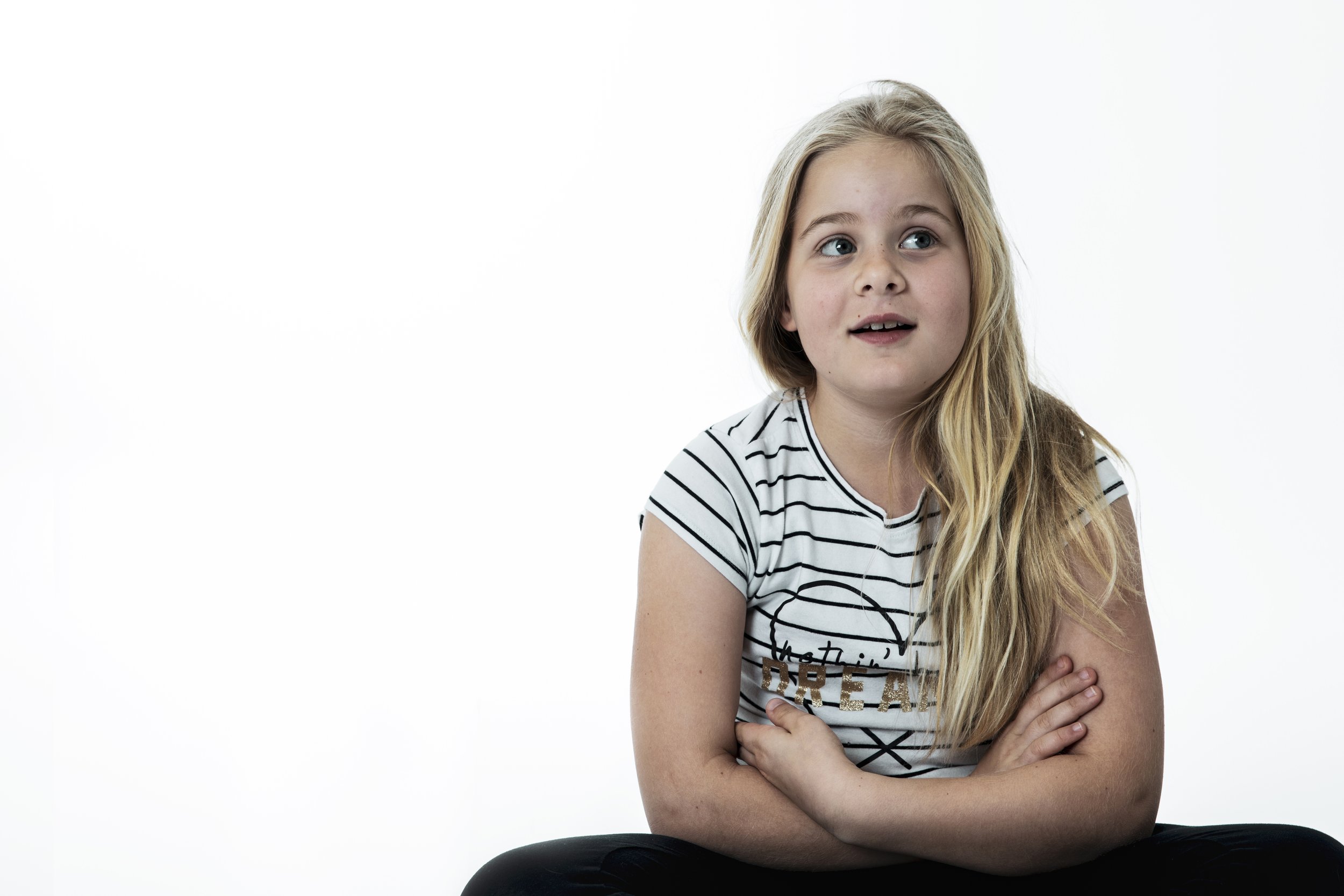 A young girl with long blonde hair, wearing a white striped t-shirt with a graphic design, sitting cross-legged against a white background, looking to the side with a curious expression.