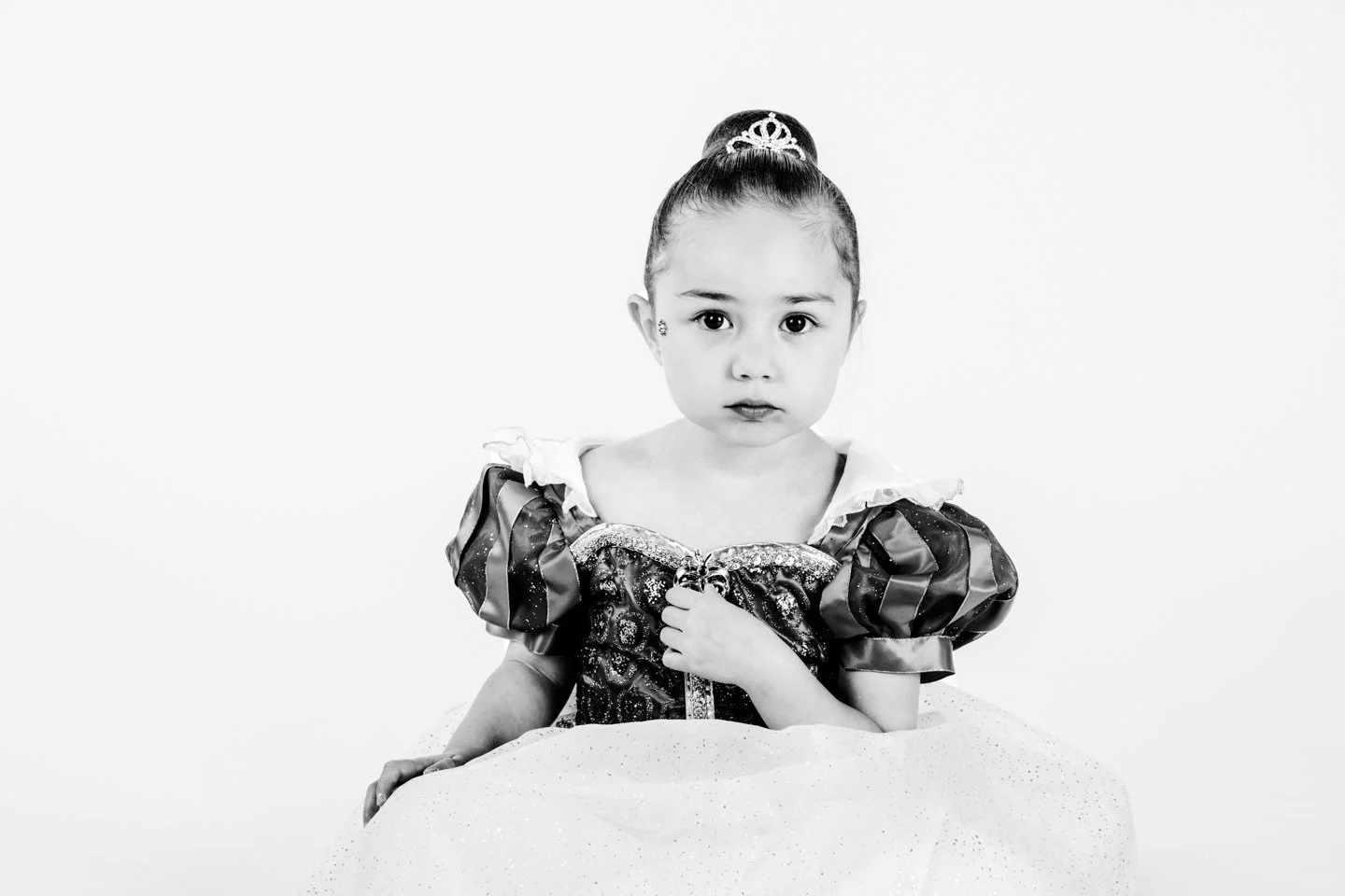 Black and white photo of a young girl dressed as a princess, wearing a tiara, with her hand on her chest and looking directly at the camera.