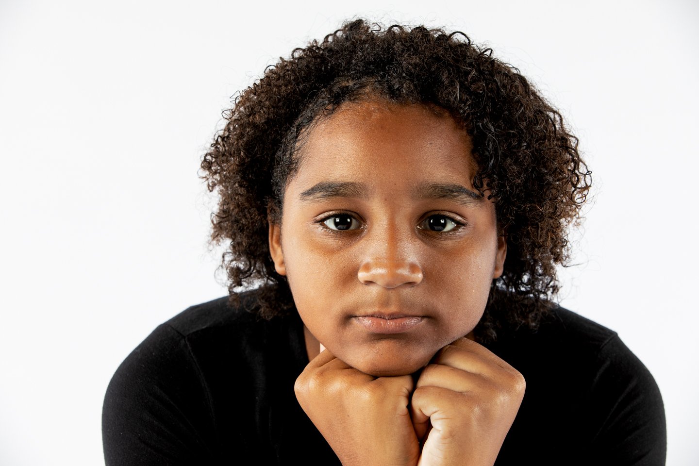 Portrait of a young girl with curly hair and a serious expression, resting her chin on her fists against a plain white background.