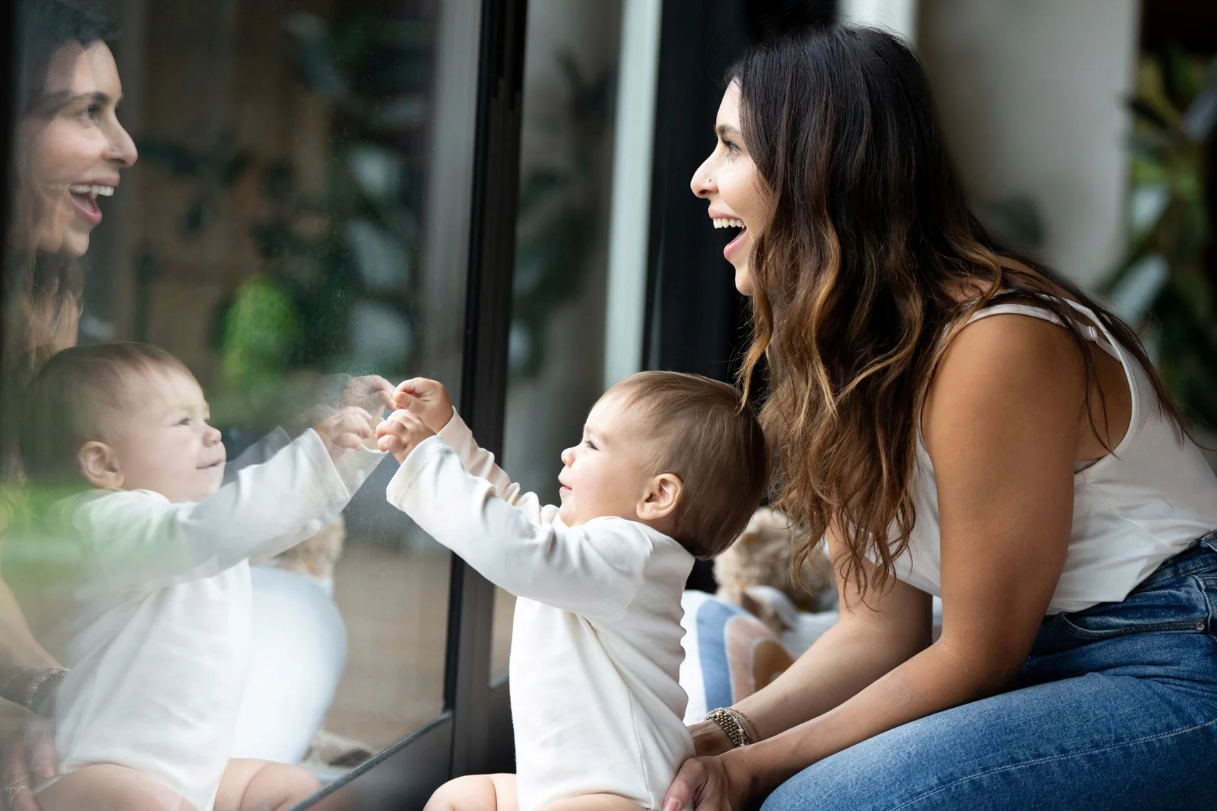 A woman and two babies smiling and touching hands through a glass door, with their reflections visible on the glass.