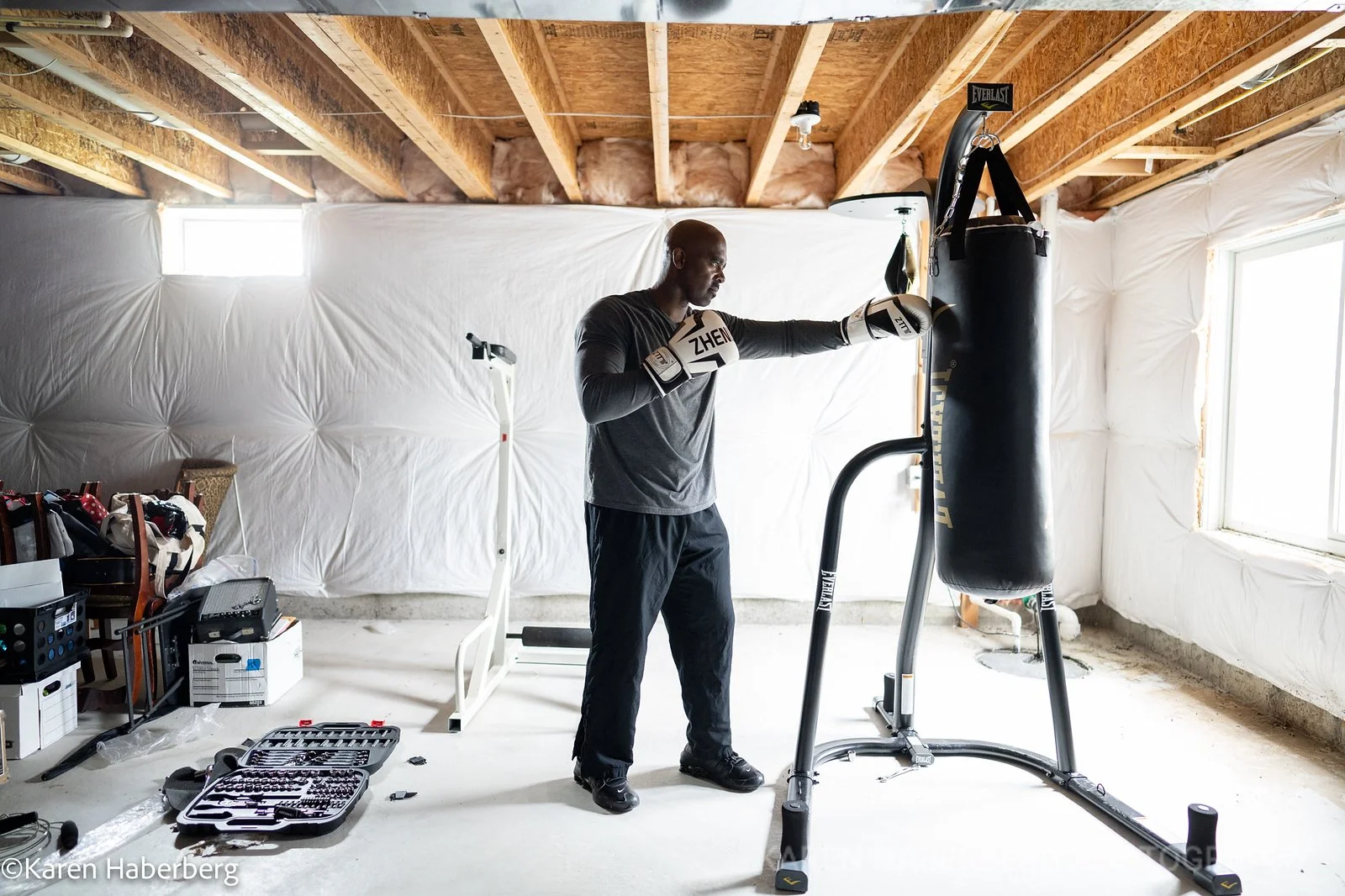 A man wearing boxing gloves practicing punches on a heavy punching bag in a basement gym. The room is unfinished, with exposed wooden beams and white insulation on the walls. There are boxes and a toolbox on the floor, and a treadmill behind him.