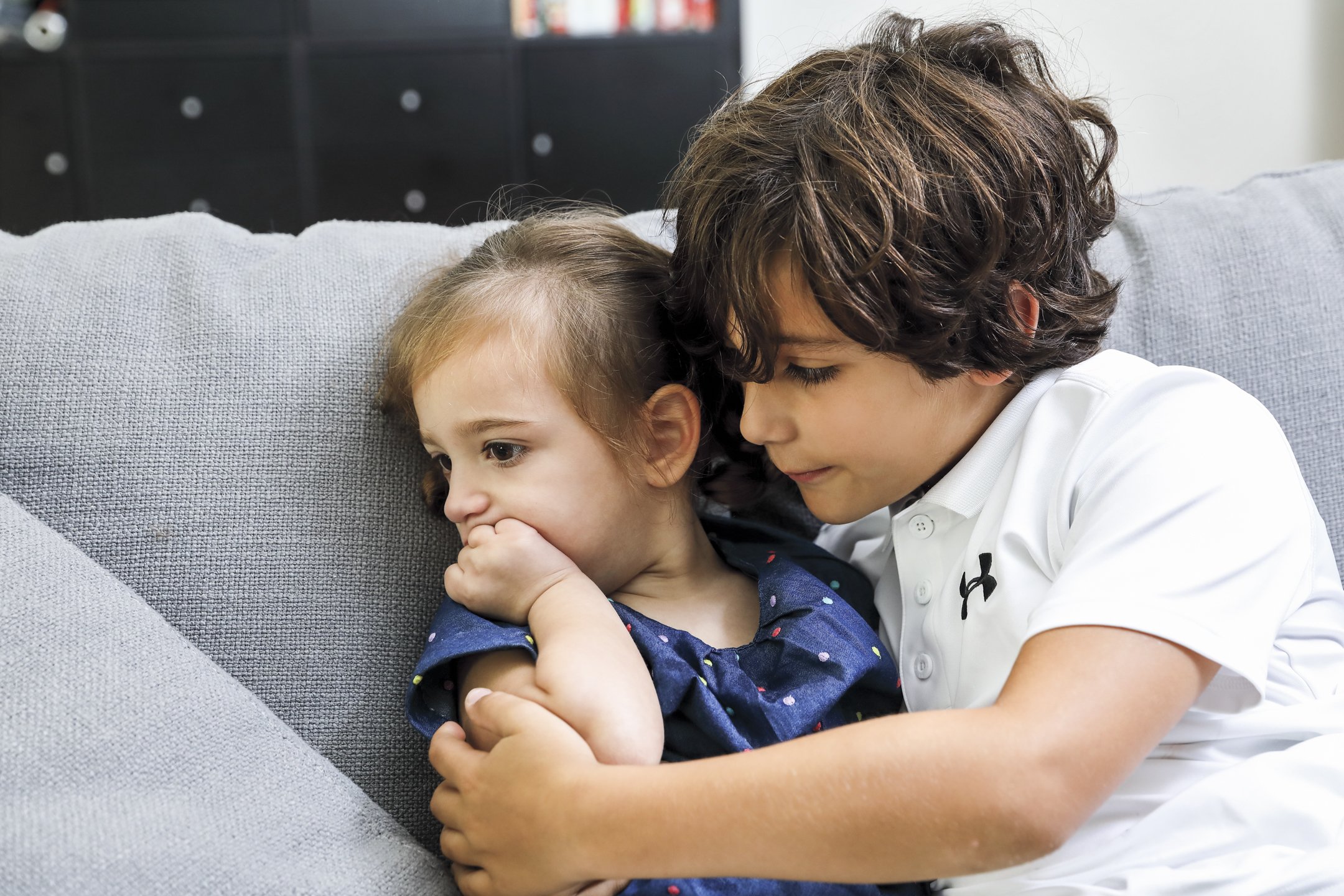 A boy comforting a girl on a gray sofa in a living room.