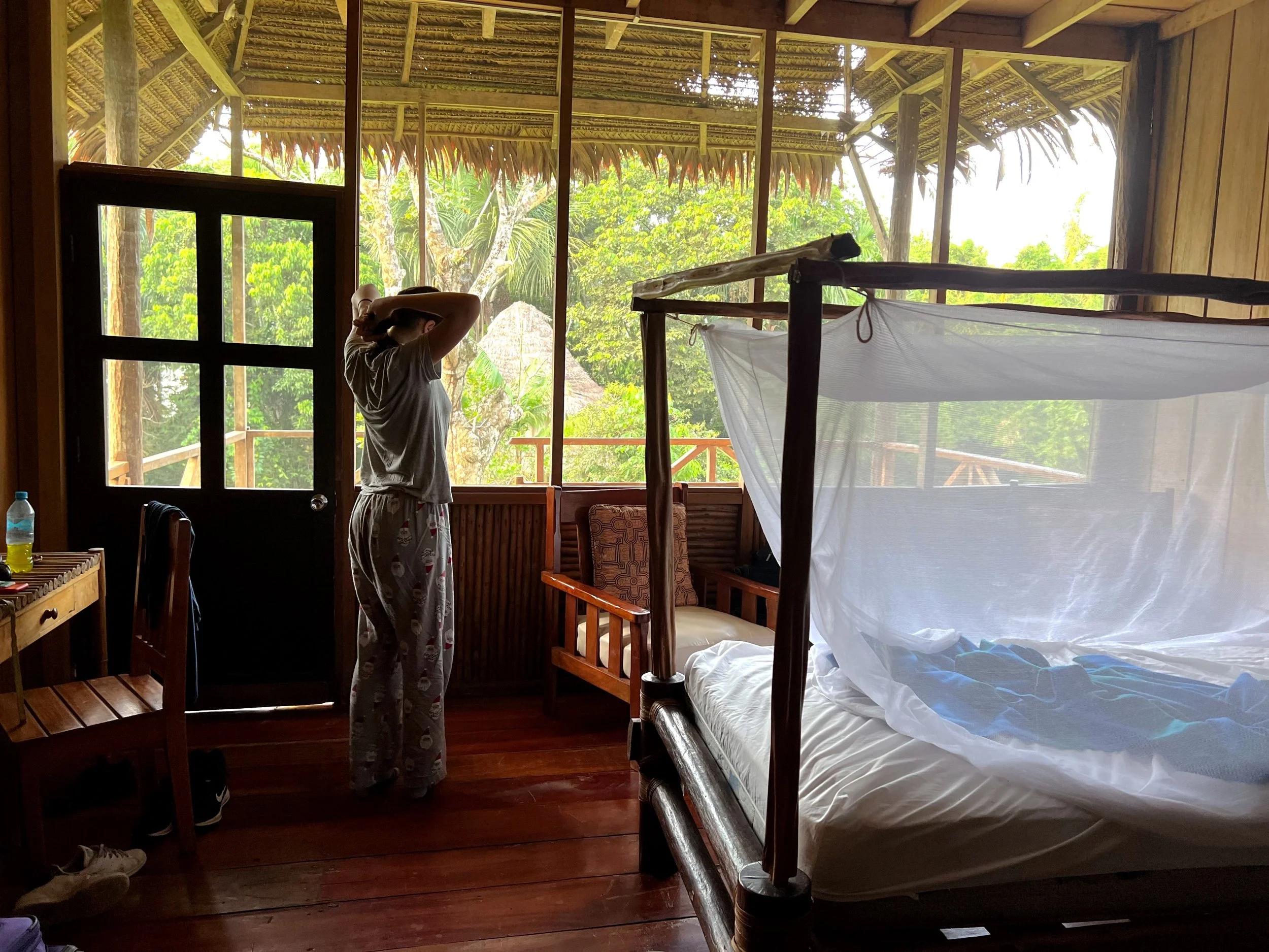 A person standing inside a wooden bungalow, looking out through large windows at lush green trees outside. The person is wearing pajamas and holding their head. There is a bed with a mosquito net in the room, and a door with a bottle on a table nearb