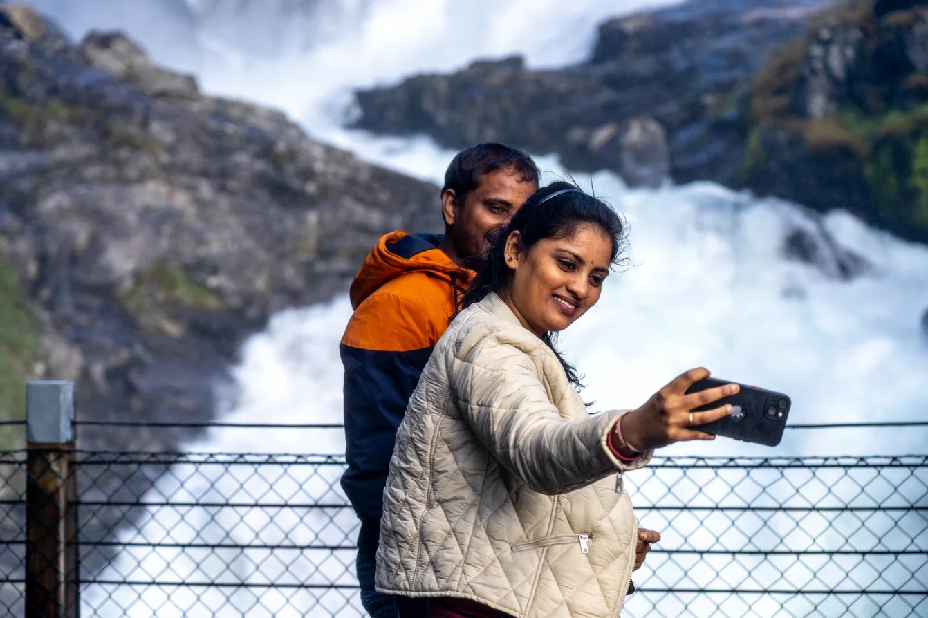 Two people, a man and a woman, taking a selfie in front of a waterfall, with the woman holding a smartphone and smiling.