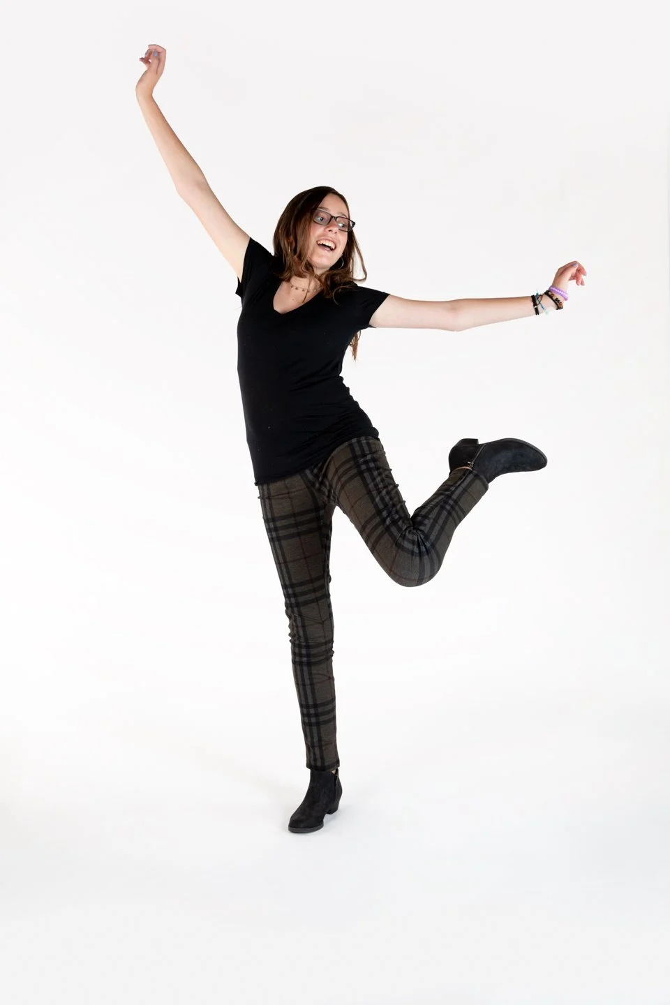 A young woman with glasses, wearing a black t-shirt, plaid pants, and boots, is jumping with her arms raised and a happy expression on her face against a plain white background.