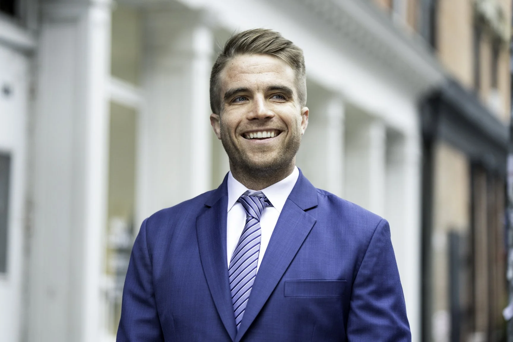 A young man with short brown hair, wearing a blue suit, white shirt, and striped tie, smiling while standing outside in an urban area.