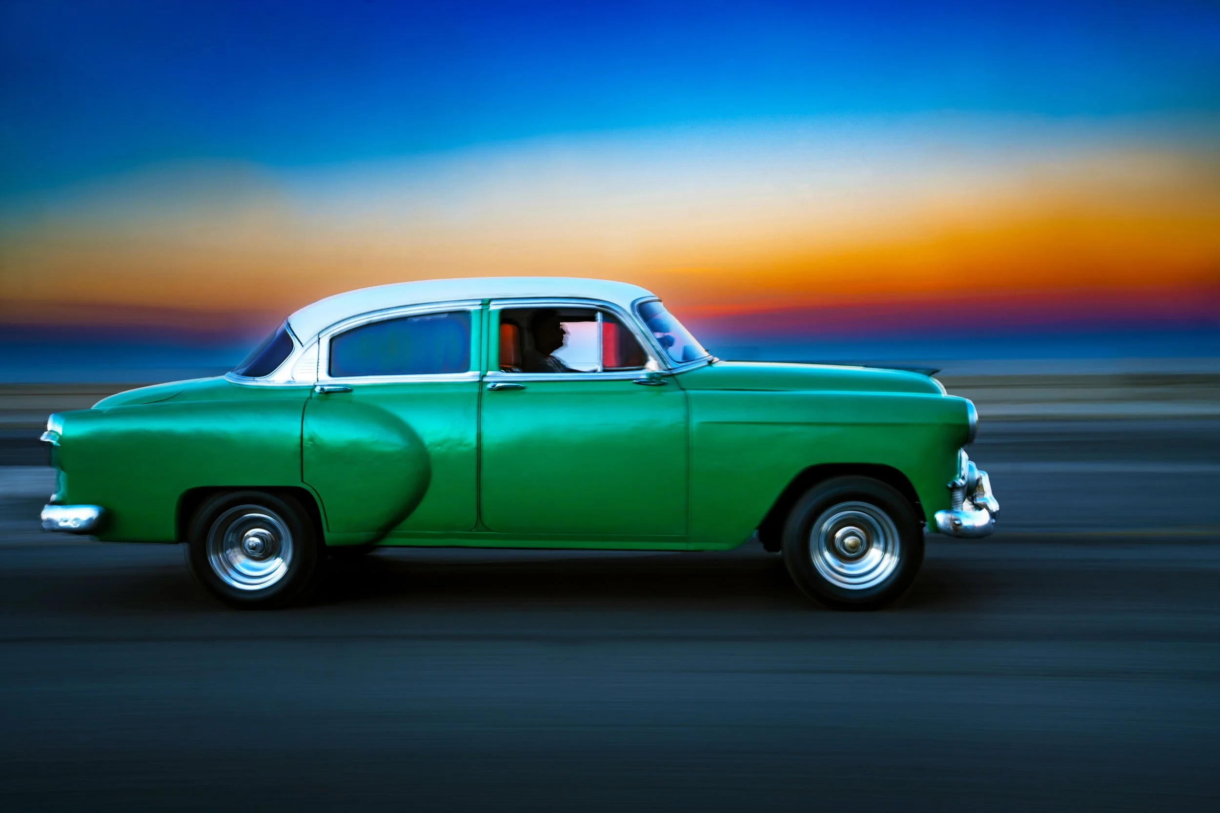 A vintage green and white car driving at sunset with a colorful sky in the background.