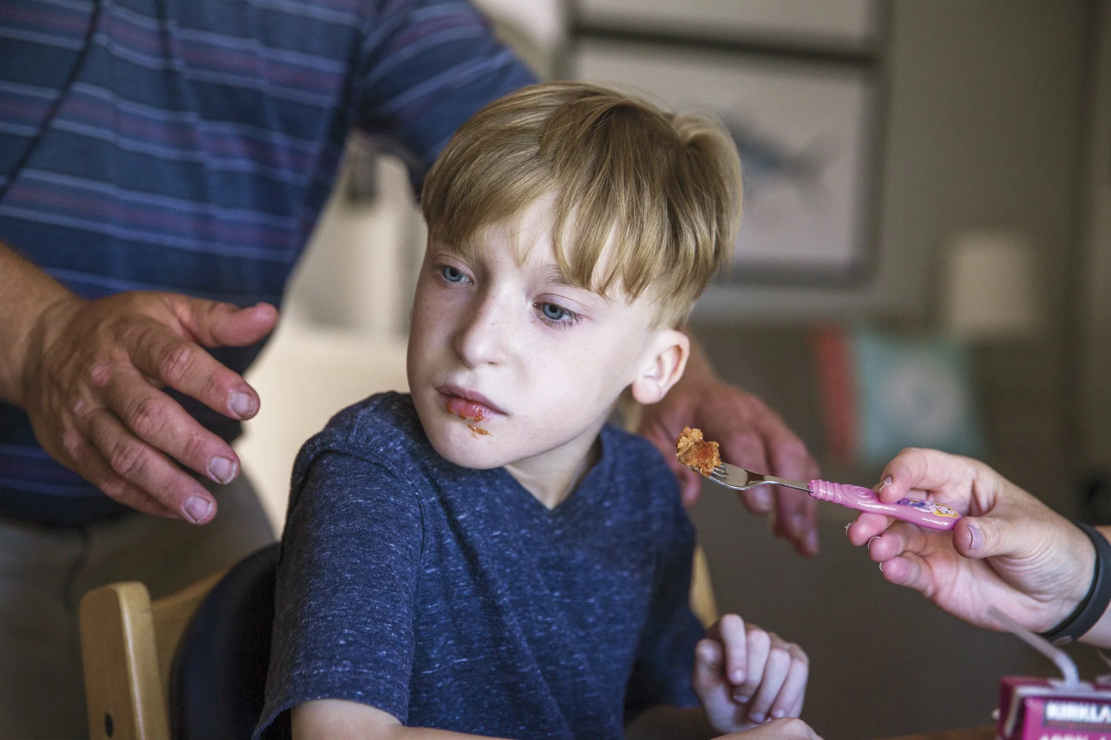 Young boy with light hair and blue eyes sitting in a high chair receiving food from a fork, with an adult hand guiding the process.