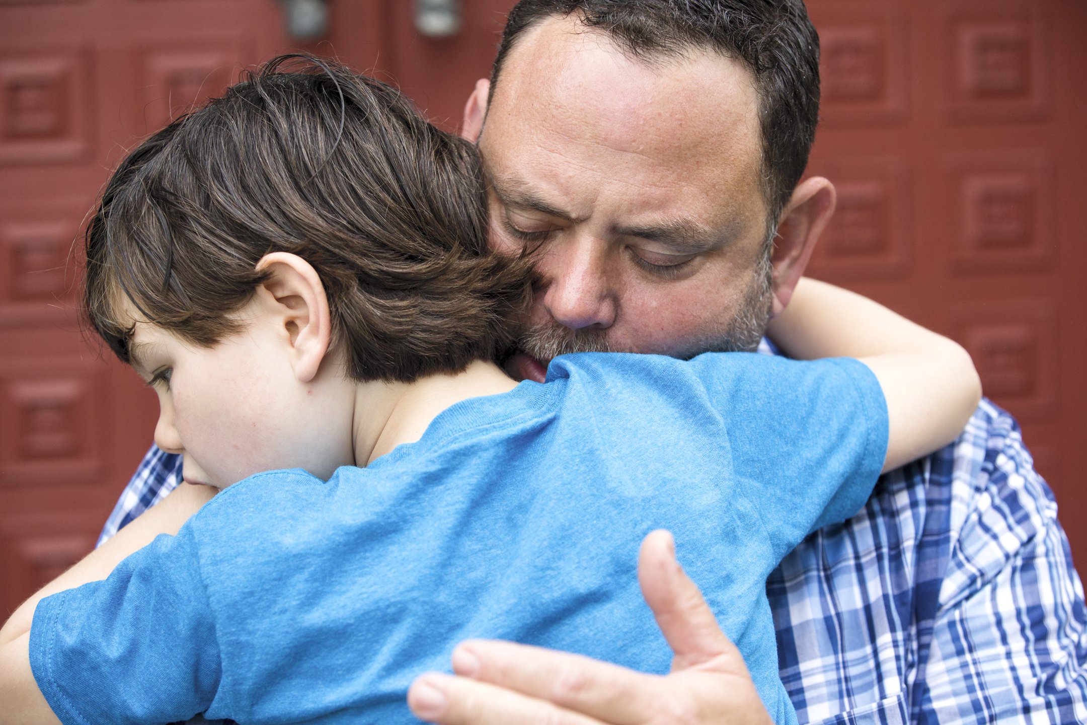 A man and a boy are hugging each other tightly with their eyes closed. The man has short dark hair and some gray, and the boy has medium-length brown hair. They are standing in front of a red wooden background.