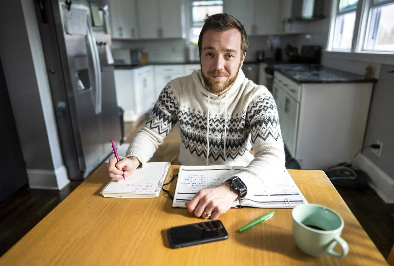 A man with a beard sitting at a wooden table, holding a pink pen and looking up at the camera. He has a planner or notebook open in front of him, with a green pen, a black smartphone, a cup of coffee, and some papers or notes. The kitchen background 