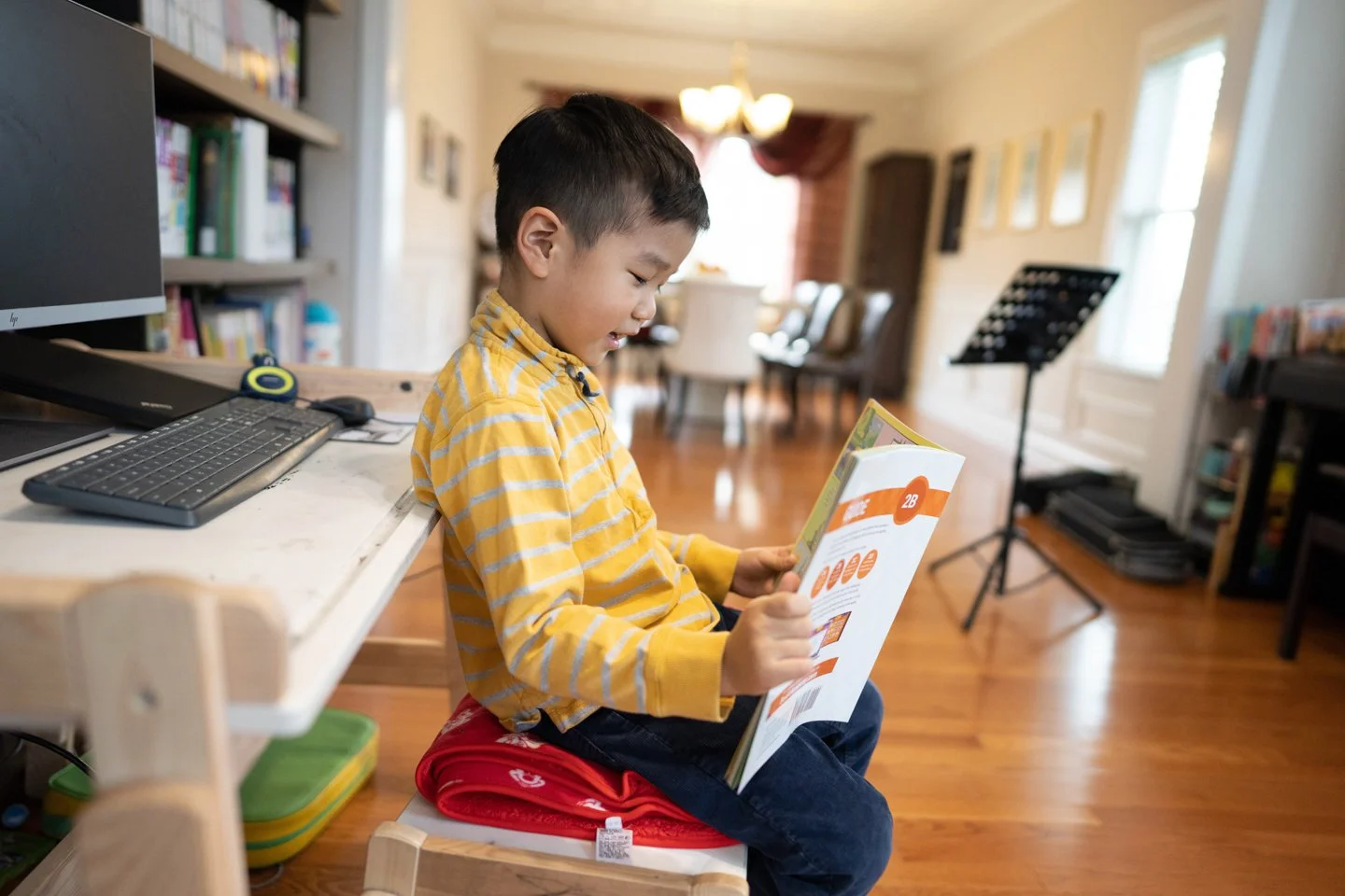 A young boy sitting on a red cushion on a wooden chair, reading a book in a cozy room with hardwood floors, a music stand, some chairs, and a bookshelf in the background.