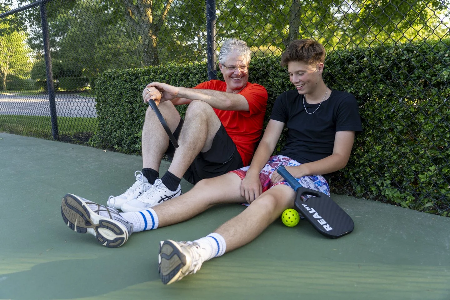 An older man and a teenage boy sitting on the tennis court, smiling and laughing, after playing pickleball. The man is wearing a red shirt, black shorts, and white shoes. The boy is wearing a black shirt, colorful shorts, and white shoes, with a pick