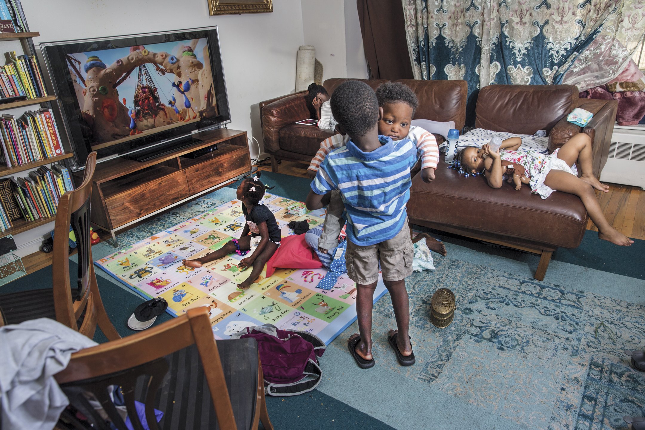 Children playing and relaxing in a living room with a large TV, bookshelves, and couches. One child is sitting on the floor on a colorful alphabet mat, while others are on the couches or standing nearby.