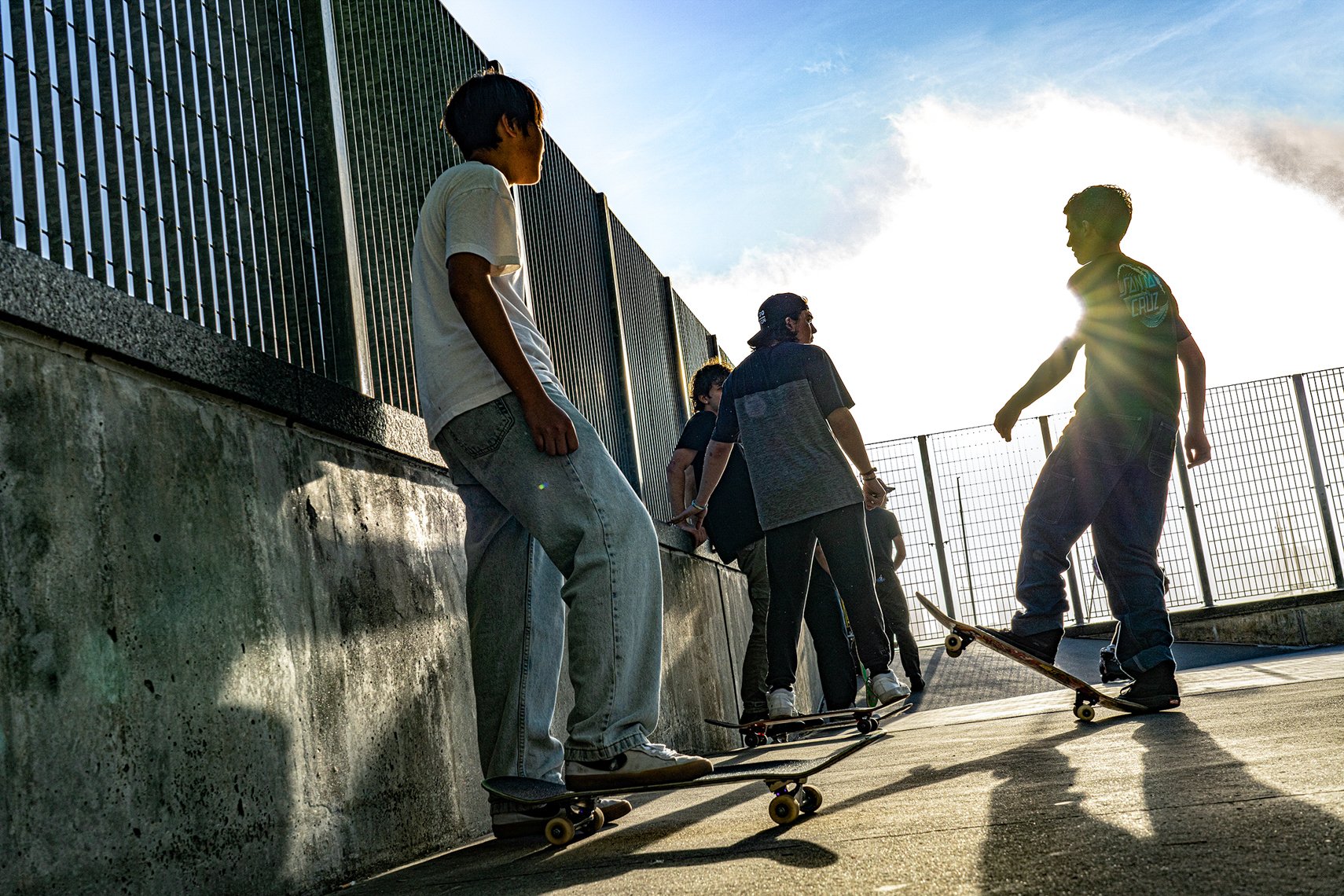 A group of young skateboarders hanging out at a skatepark during sunset, with some sitting on the ledge and others preparing to skate.
