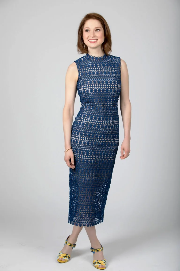 A woman with shoulder-length brown hair wearing a sleeveless, floor-length, dark blue lace dress and floral high heels, smiling and posing against a light gray background.