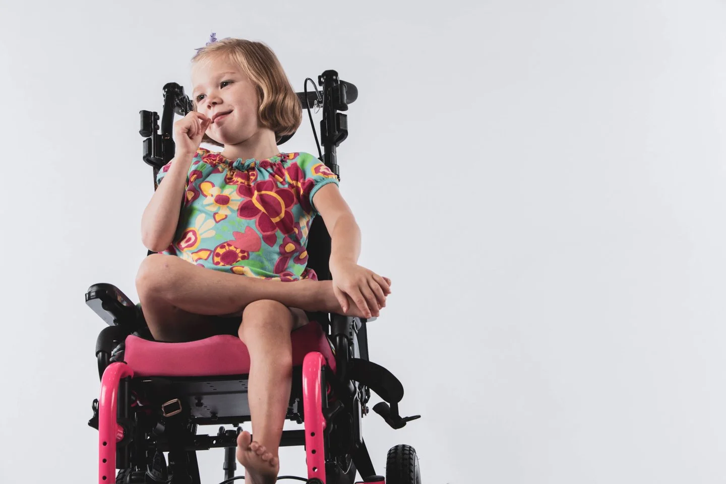 A young girl with blonde hair sitting cross-legged in a black and pink wheelchair, wearing a colorful floral dress, with her finger on her lips, looking to the side, against a plain light gray background.