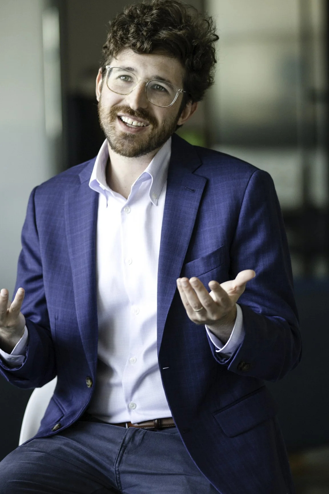 A man with curly brown hair and a beard, wearing glasses, a white shirt, and a blue suit, smiling and gesturing with his hands.