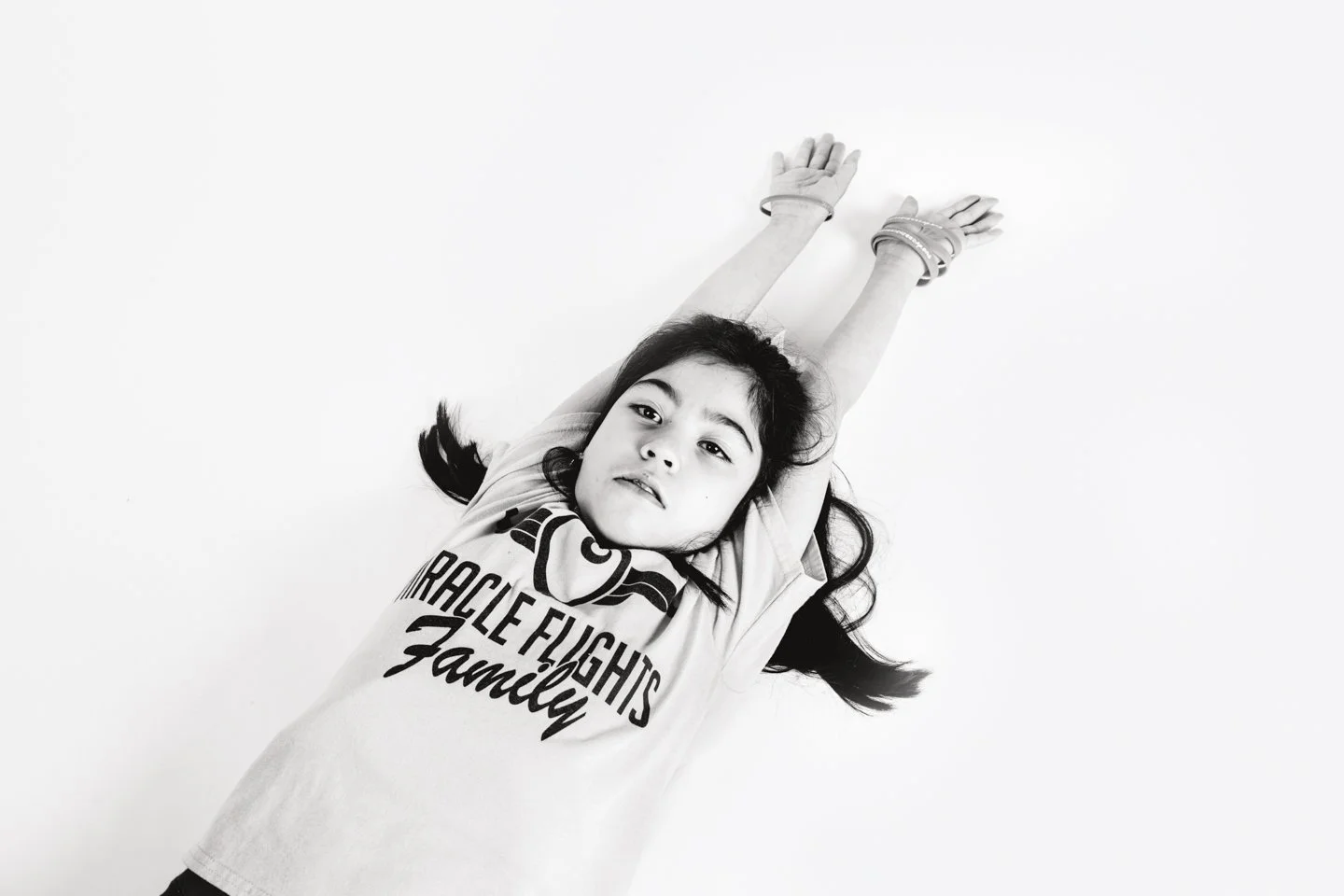 Black and white photo of a girl lying on her back with arms and legs stretched upwards, wearing a T-shirt that says 'Miracle Fights Family' and multiple bracelets on her wrists.