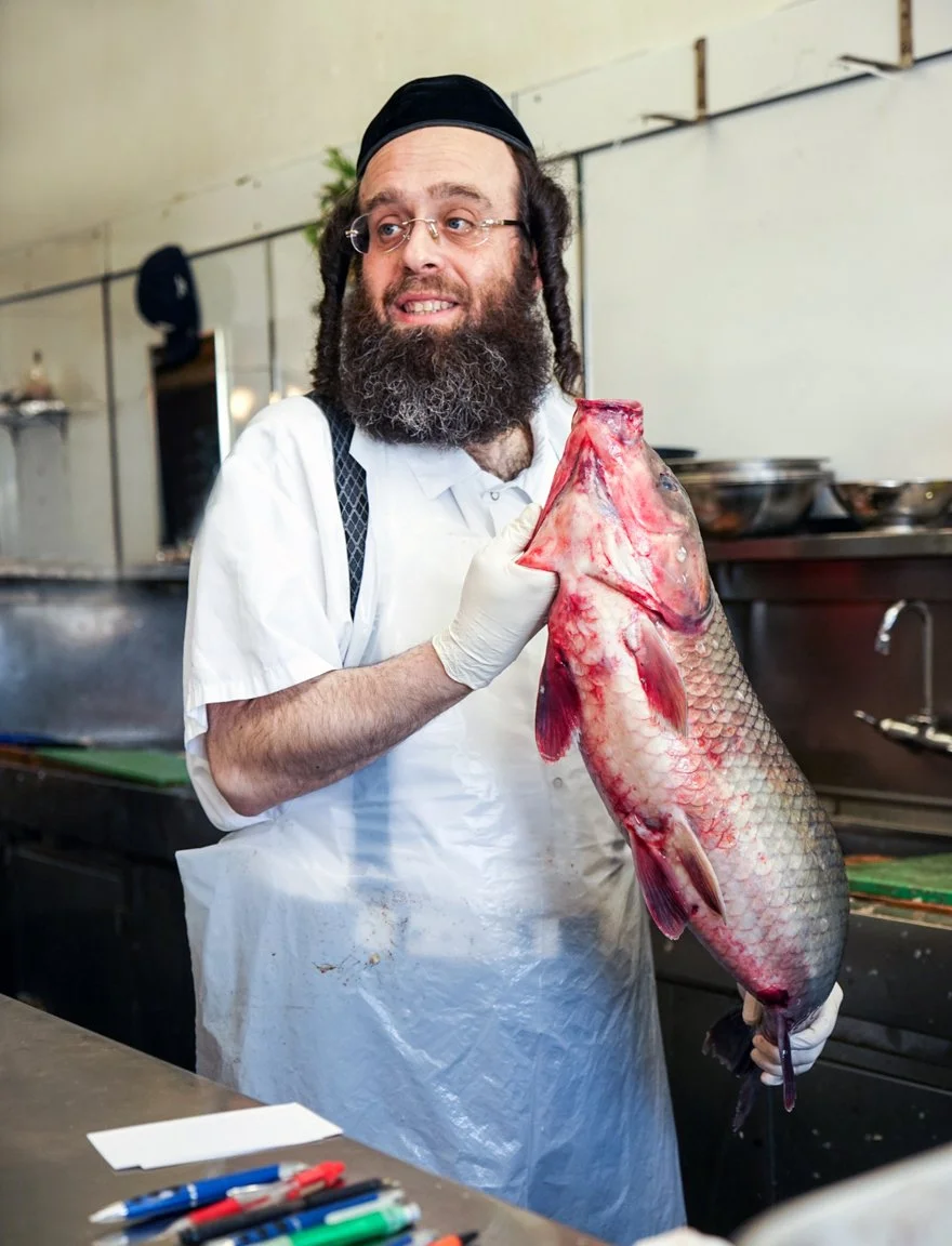 A man with glasses, a beard, and dreadlocks holding a large fish in a kitchen or fish market setting.